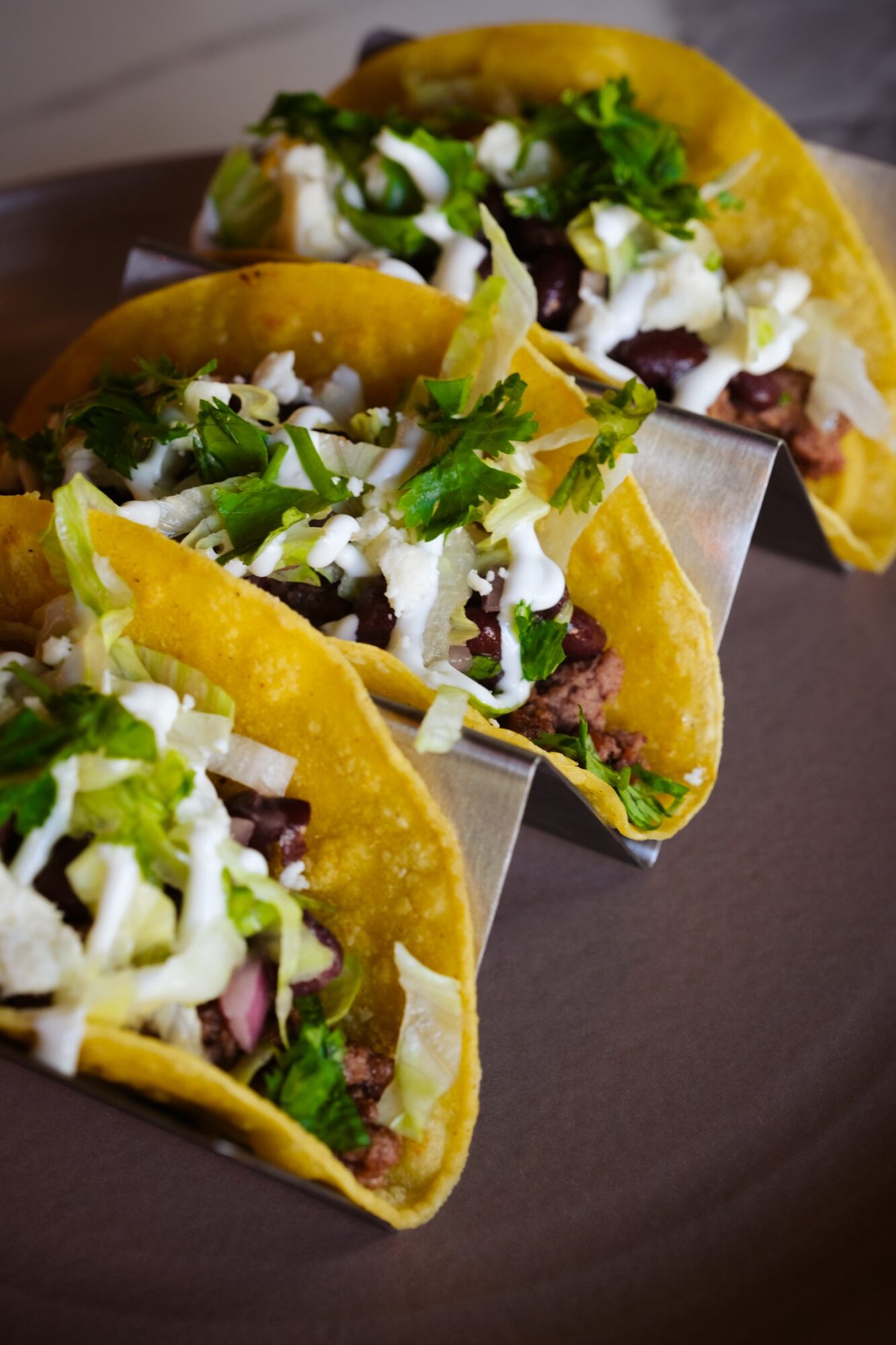 Three tacos filled with meat, lettuce, cheese, and cilantro on a dark surface.
