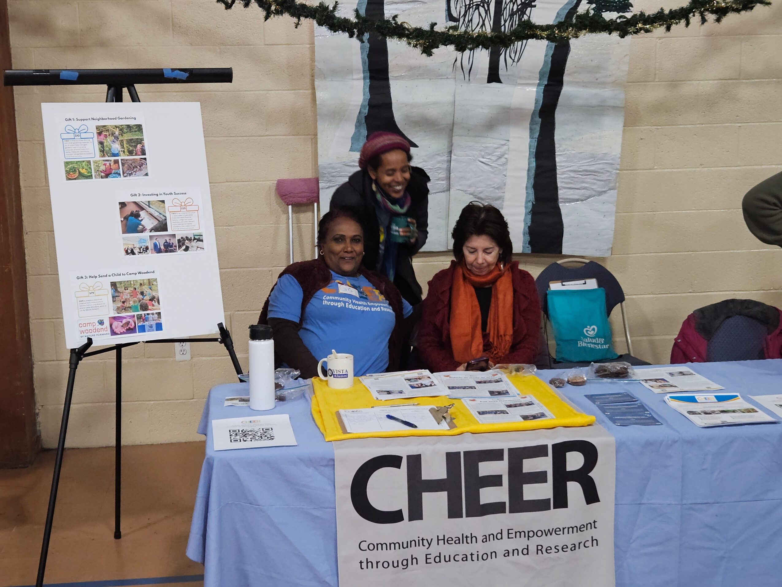 Three women sit behind a table with community health and research materials, with a large banner and display board in the background.