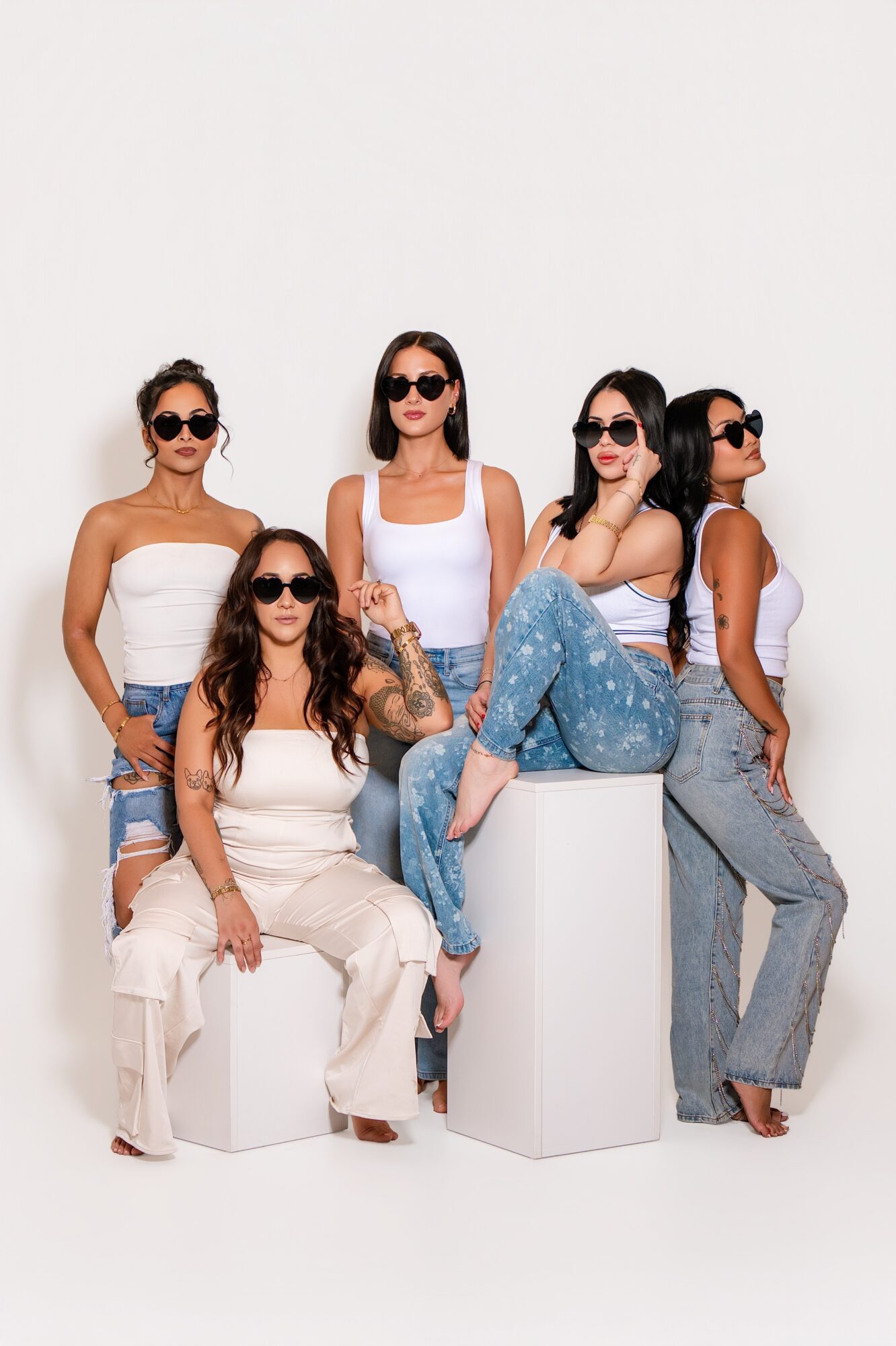 Five women with sunglasses pose against a white background, some sitting and some standing, wearing casual outfits.