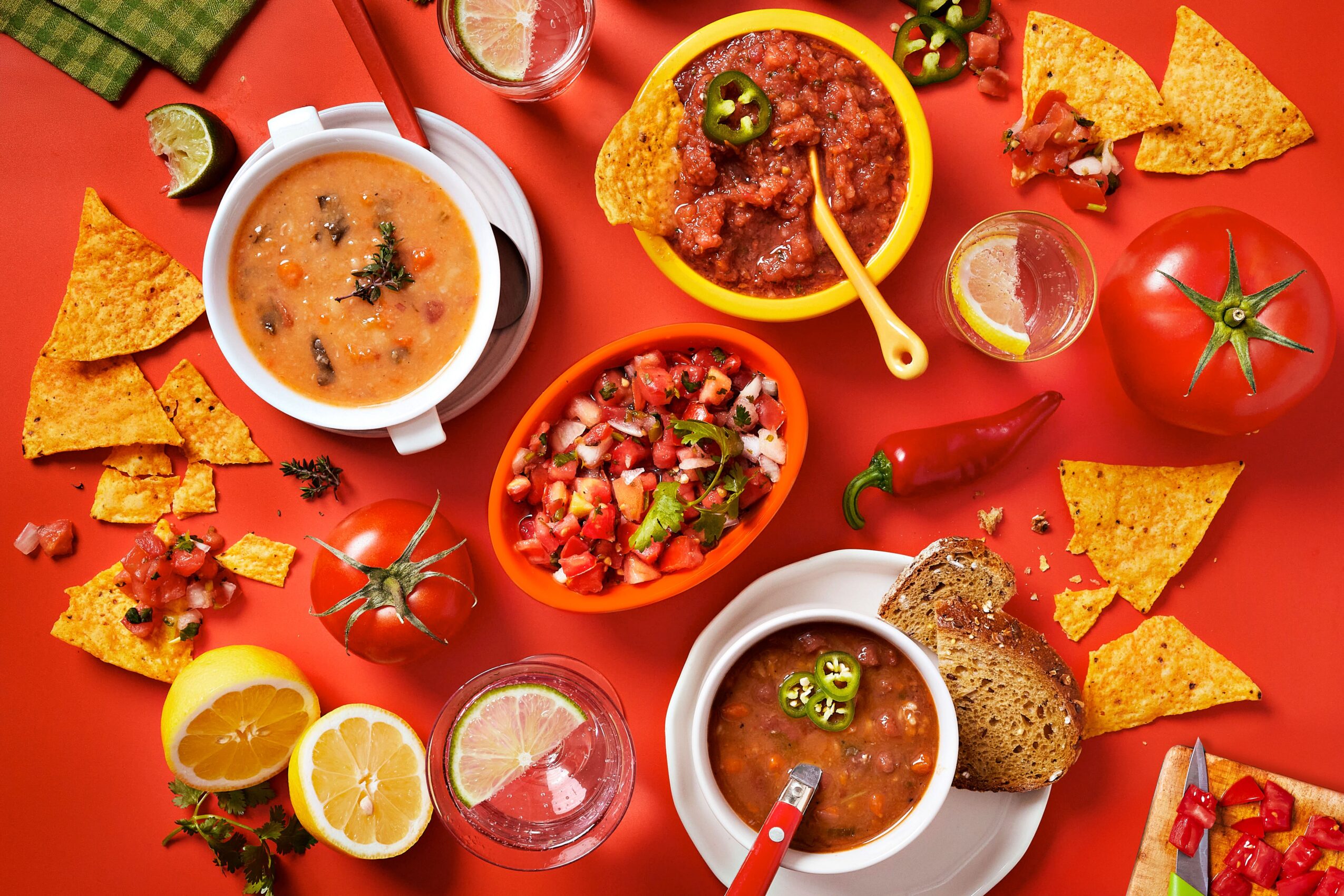 Assorted Mexican dishes including soup, salsa, chips, tomatoes, lemon, and bread on a red background.