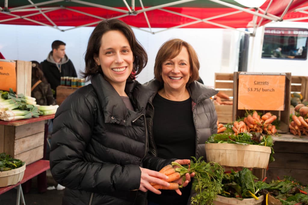 Two women smiling at a market stall with vegetables and flowers, under a red canopy.