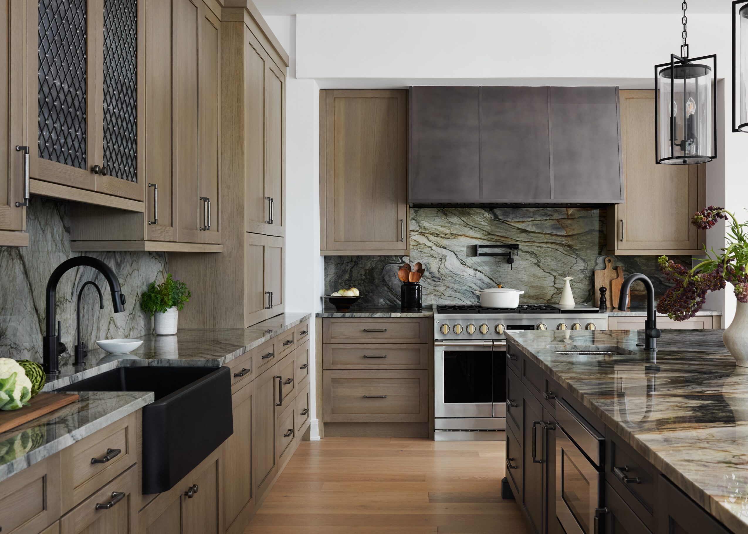 Modern kitchen with wooden cabinets, marble countertops, and a stove with a gray range hood, featuring plants and kitchen utensils.
