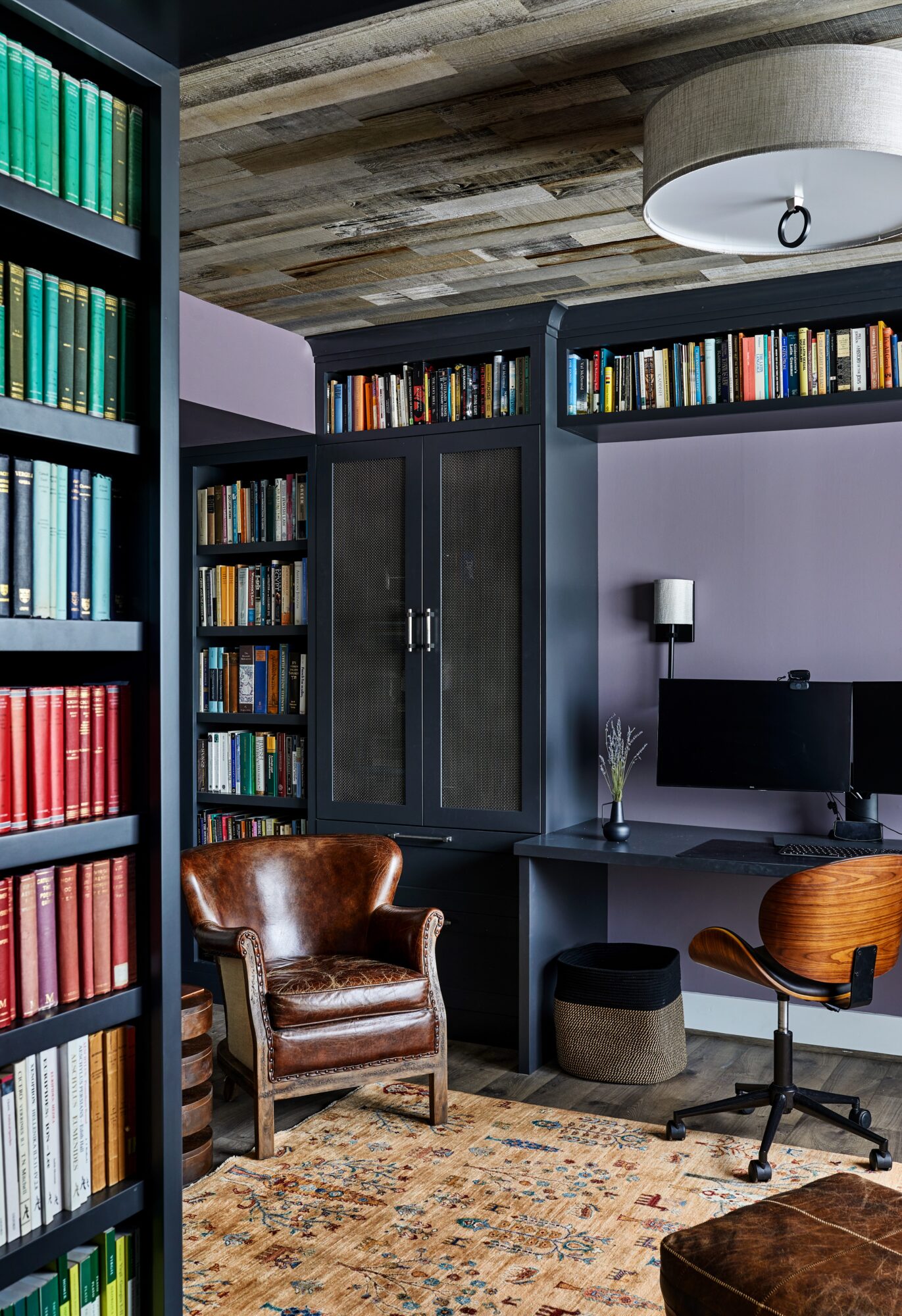 Home office with bookshelves, a leather armchair, a desk with a computer, and a wooden chair, with a patterned rug and ceiling light.