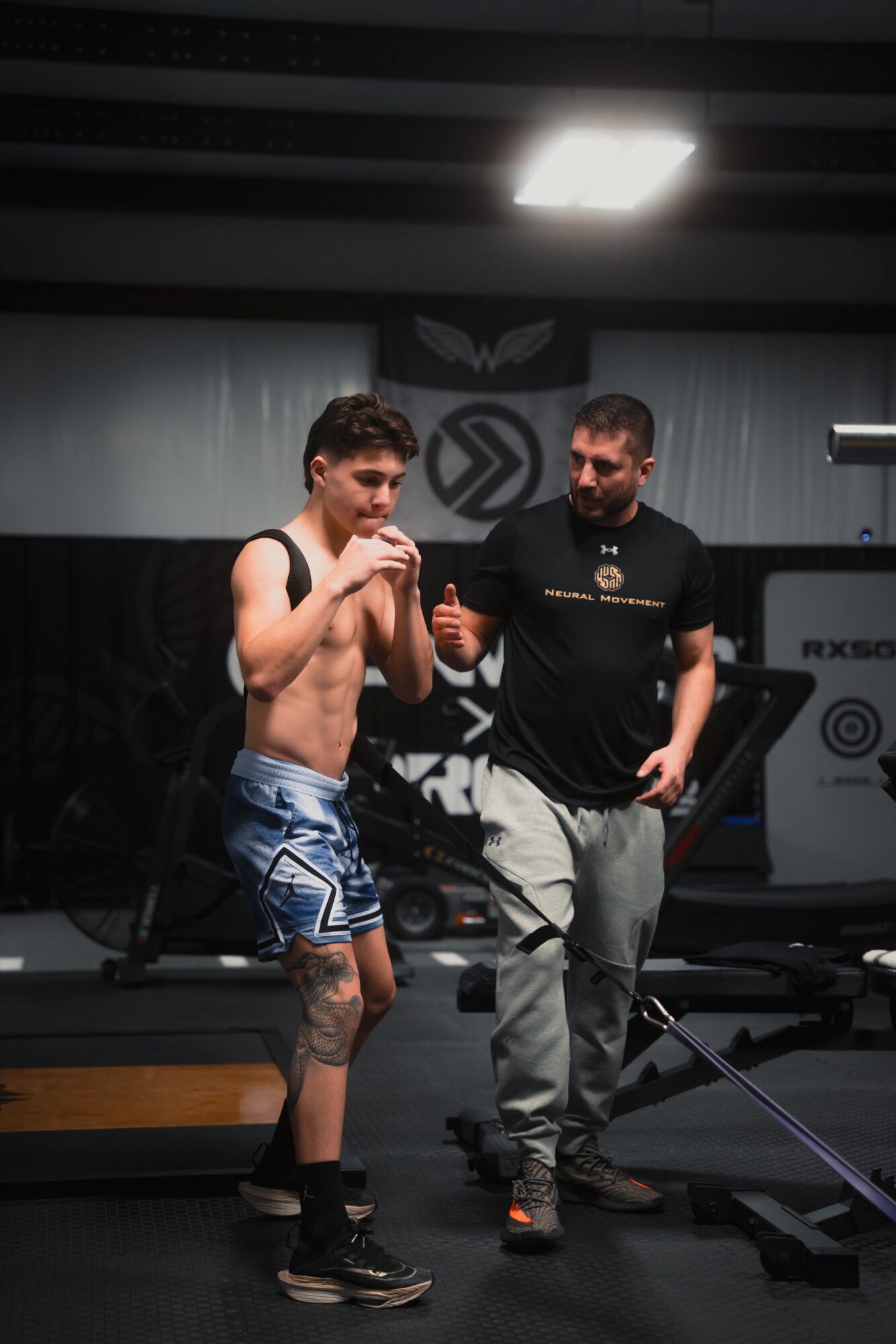 Young man with a tattoo on his left leg, shirtless, wearing shorts, talking to a trainer in a gym.