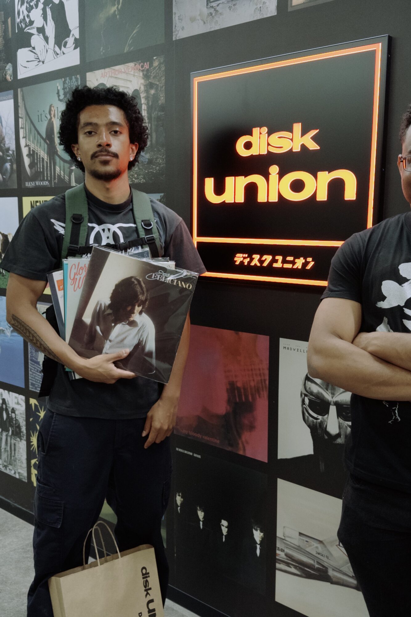 Person with curly hair holding a vinyl record, standing next to a sign that says 'disk union', in a store or exhibit.