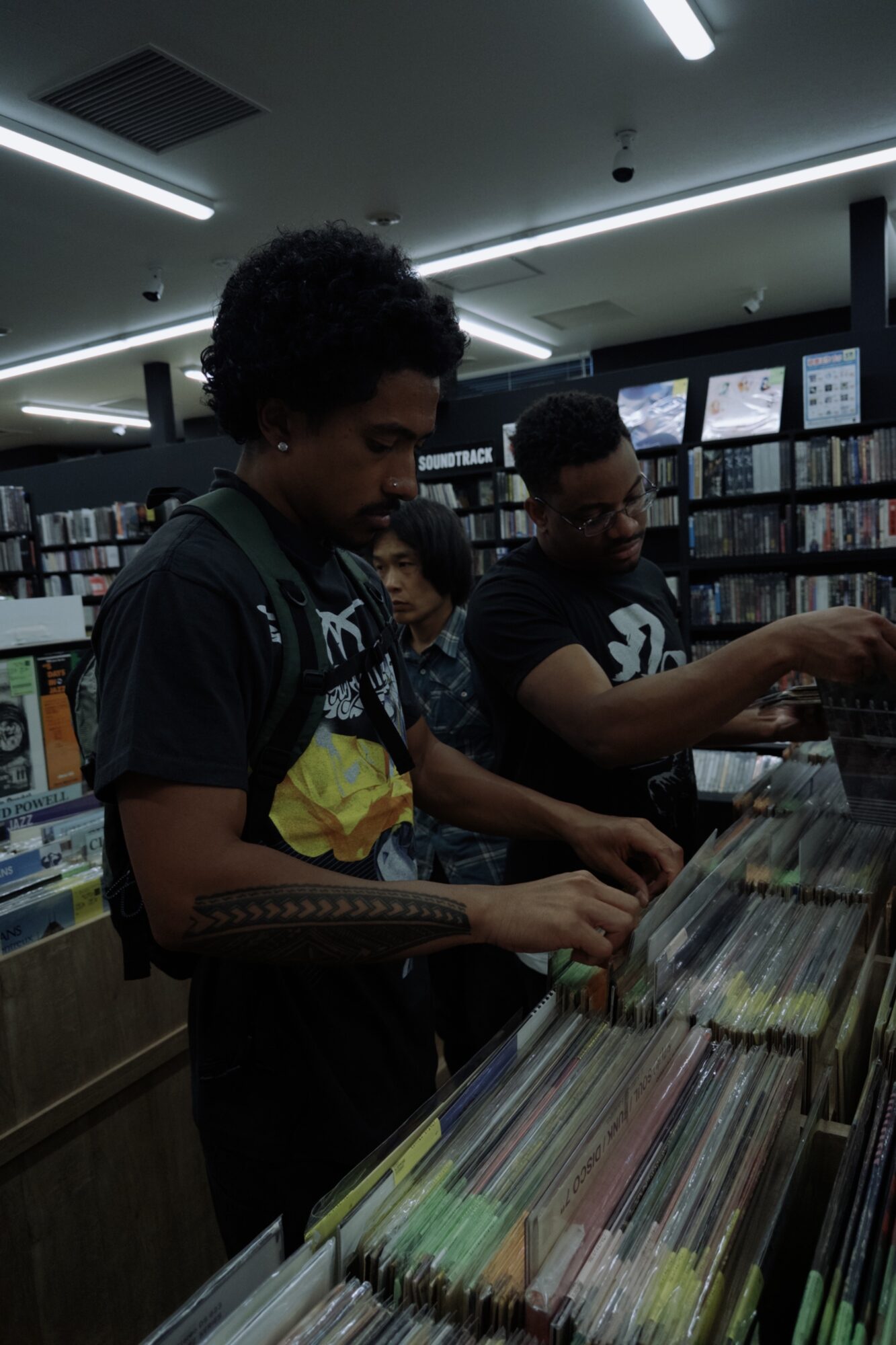 Three people browsing records in a store with shelves filled with vinyls, illuminated by ceiling lights.