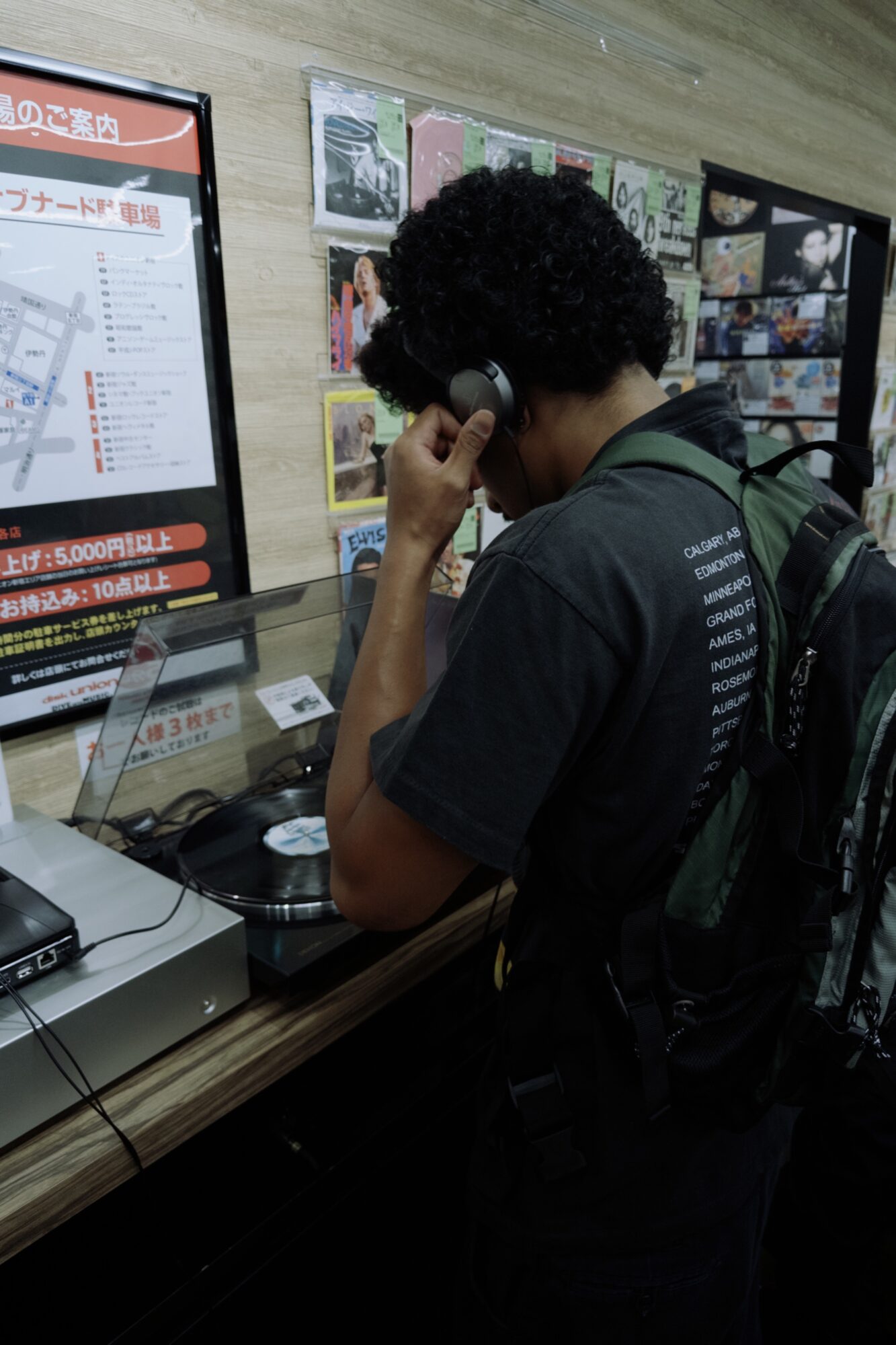 Person with curly hair using a phone at a counter with a backpack, surrounded by posters and a display board.