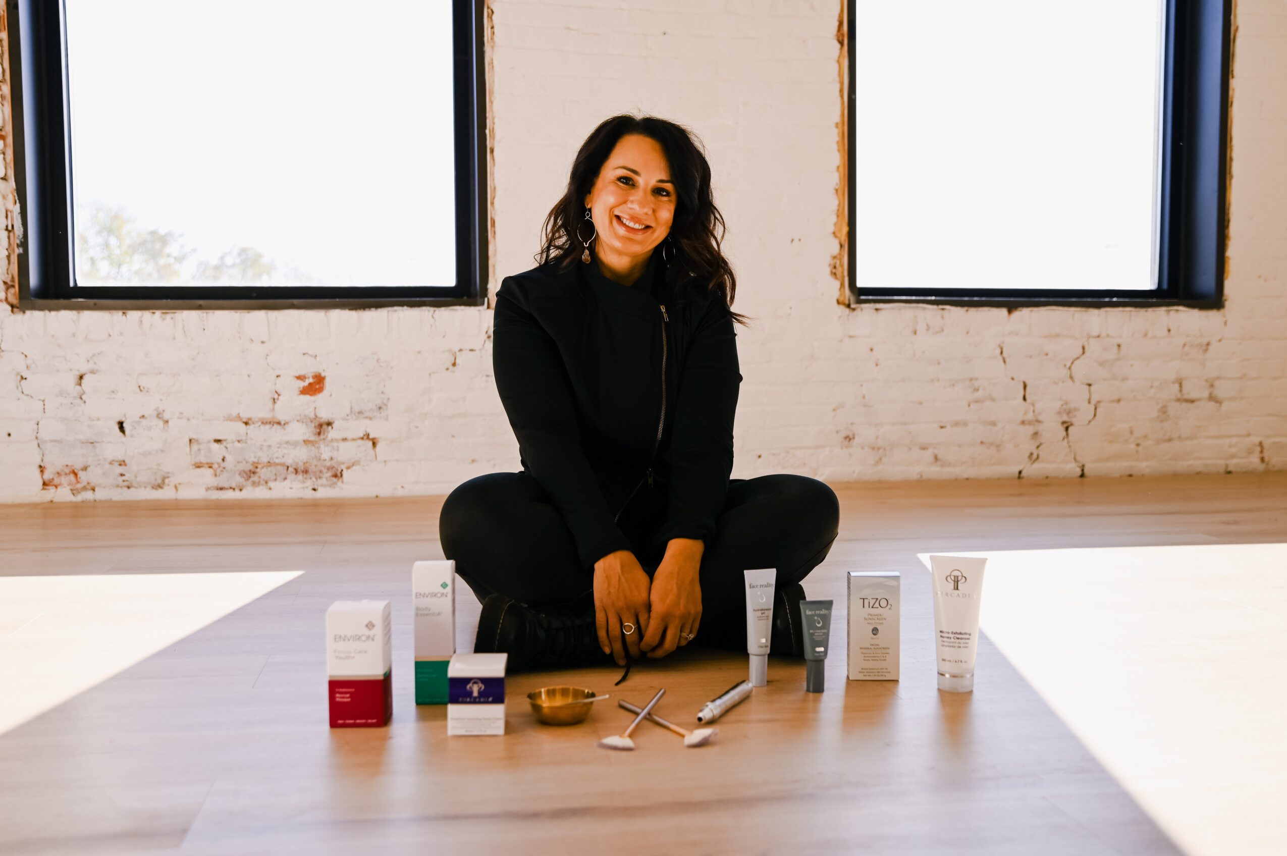 Woman sitting cross-legged on floor in front of skincare products, smiling, with large windows behind her.