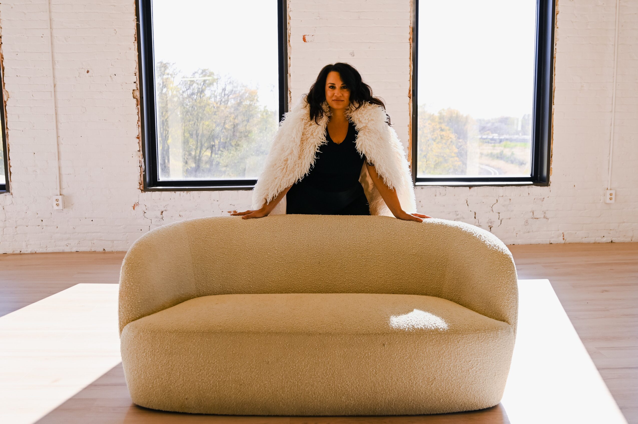 Woman standing behind a beige sofa in a bright room with large windows, sunlight streaming in.
