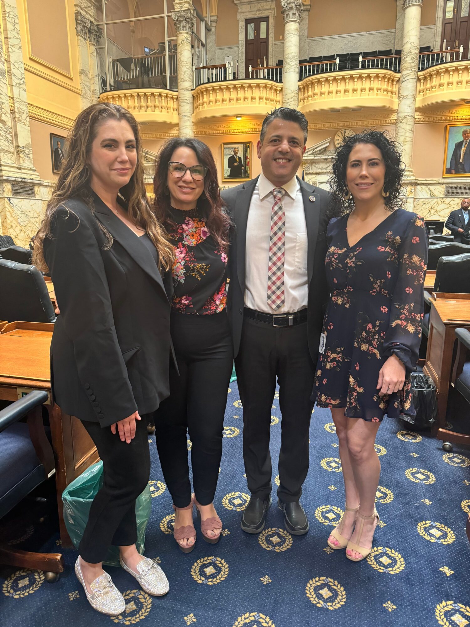 Four people standing in a room with ornate decor, dressed in formal and semi-formal attire, smiling at the camera.