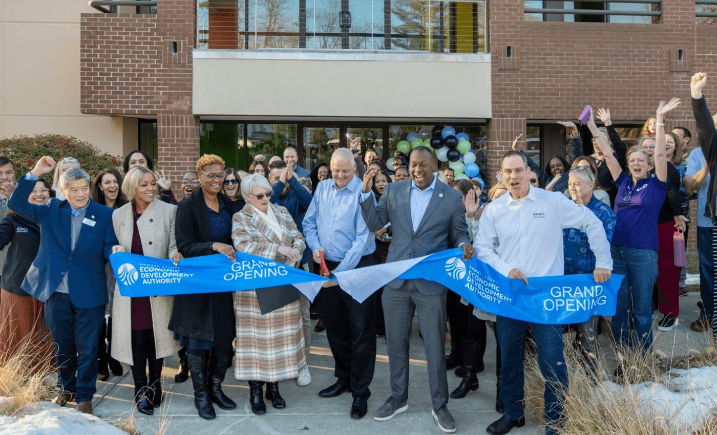 Group of people celebrating grand opening with ribbon cutting outside building, some raising hands, smiling, and holding scissors.