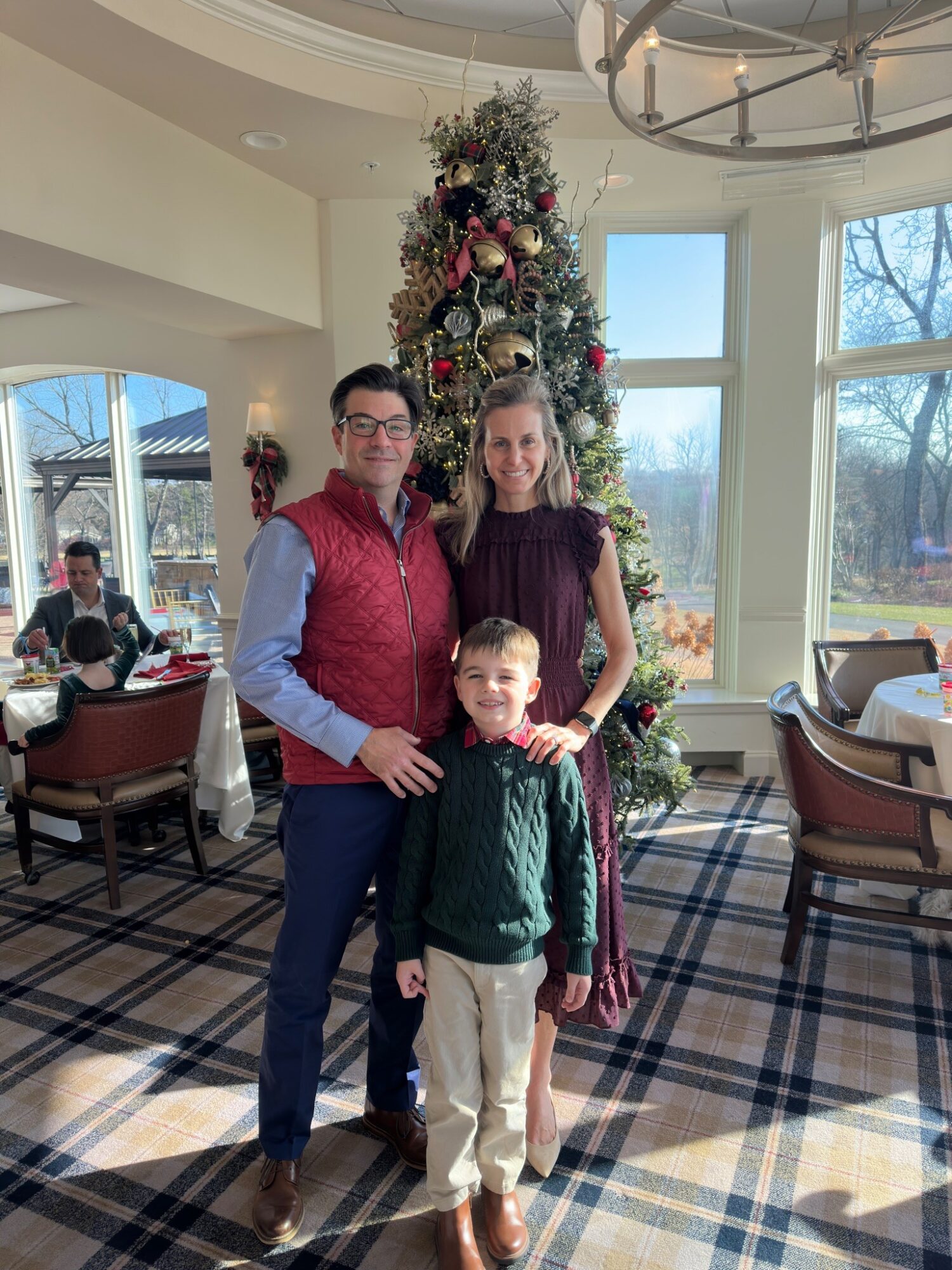 Three people, two adults and a child, stand in front of a decorated Christmas tree inside a bright room with large windows.