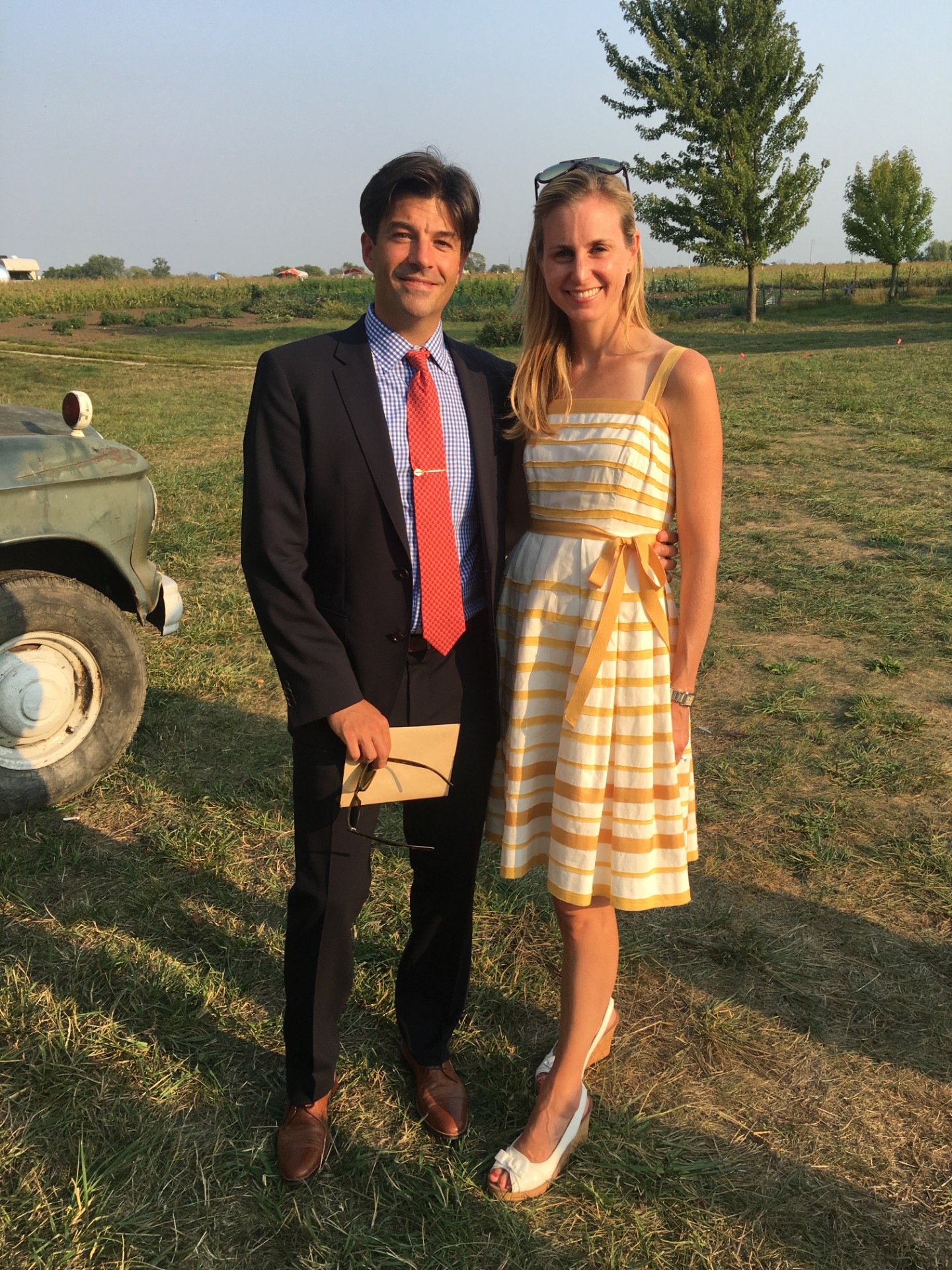 A man in a suit and a woman in a yellow striped dress stand outdoors on grass, smiling, with trees in the background.