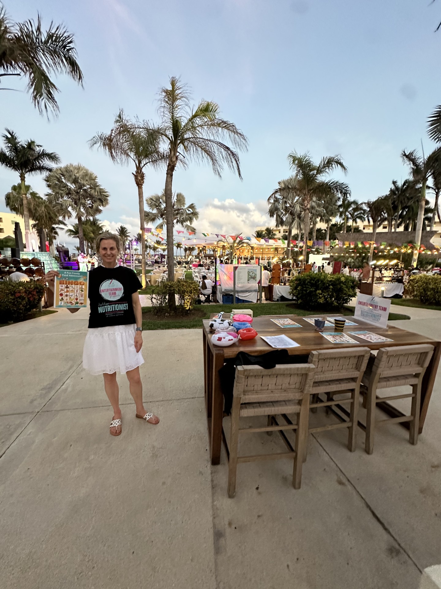 Person standing outdoors near a table with chairs, palm trees, and a beach scene in the background.