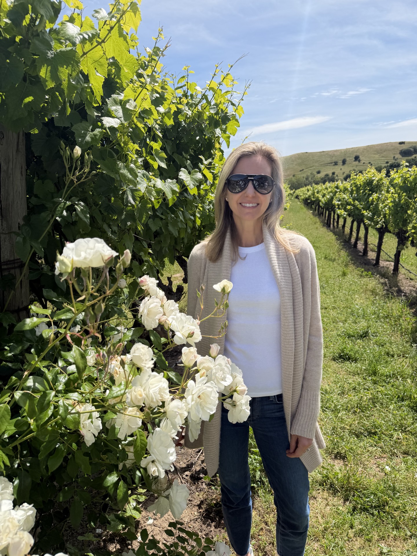 Woman with sunglasses standing in a vineyard with white flowers and green vines, sunny sky, wearing a beige cardigan and white shirt.