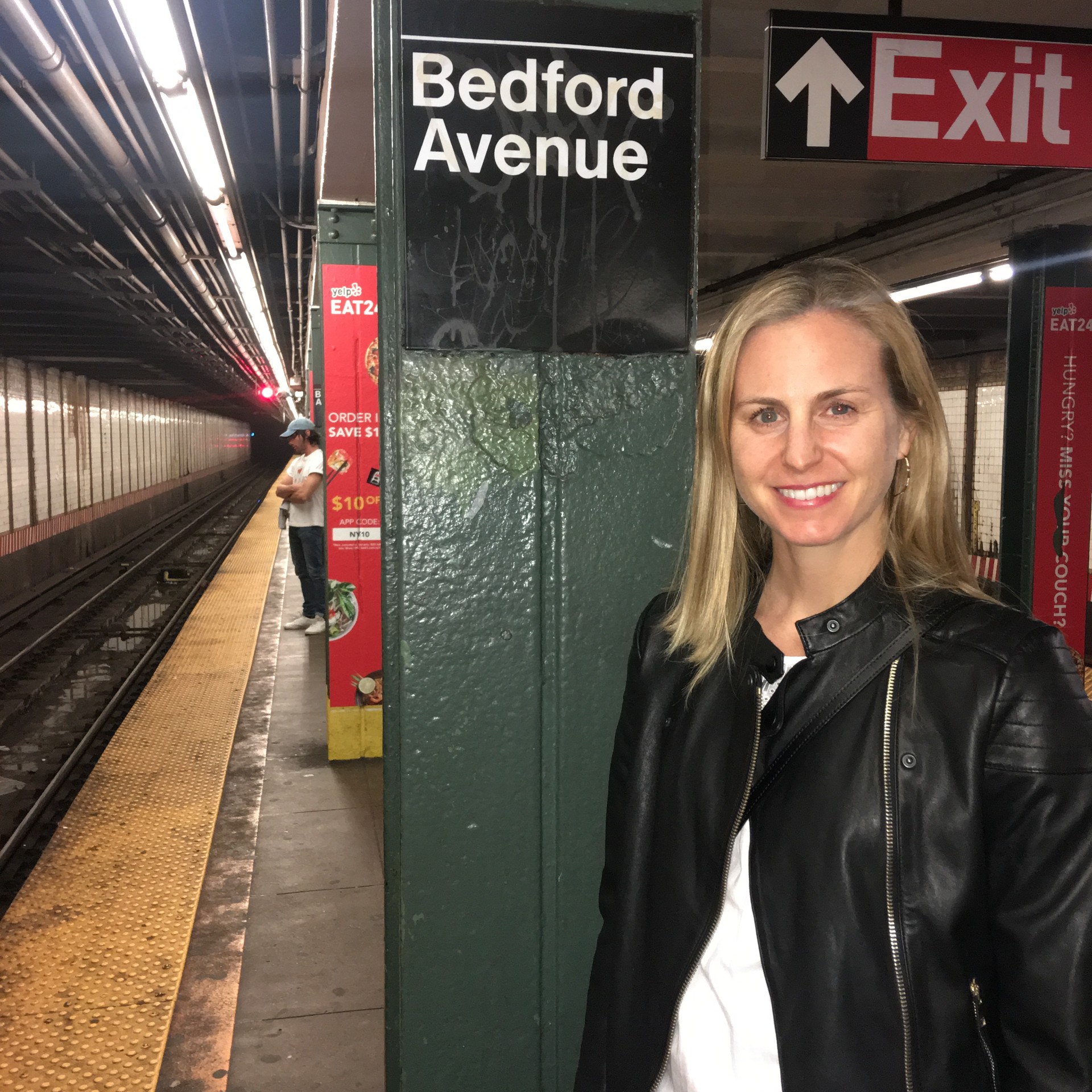 Smiling woman with blonde hair in black leather jacket on subway platform, Bedford Avenue sign visible, train tracks to the side.