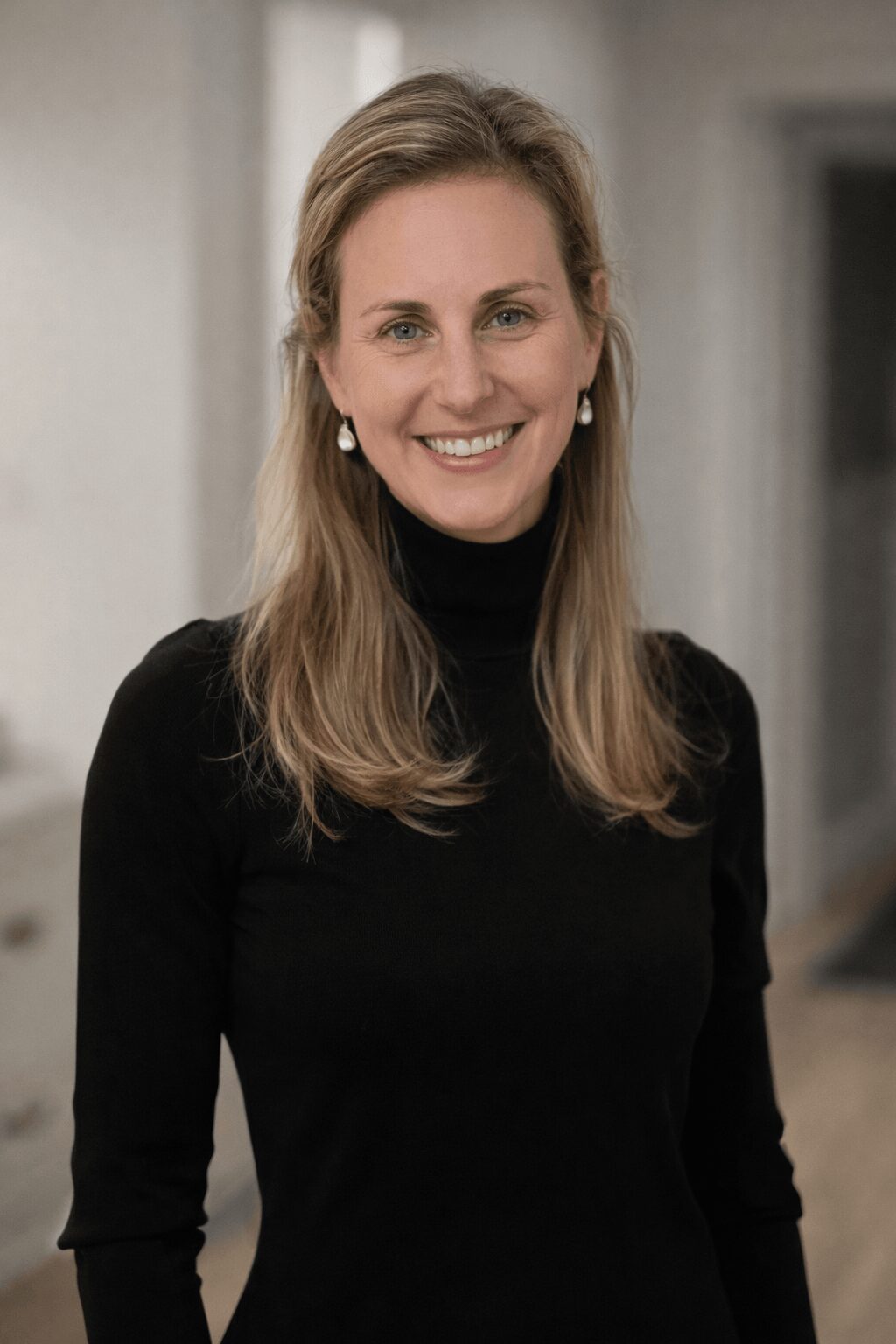Woman with long blonde hair smiling, wearing a black turtleneck, in an indoor setting.