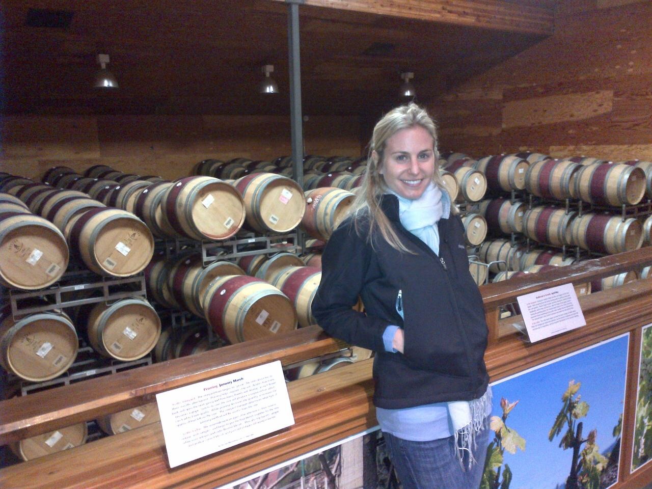 Woman with blonde hair smiling, standing in a wine cellar with stacked barrels, wearing a dark jacket and light blue scarf.