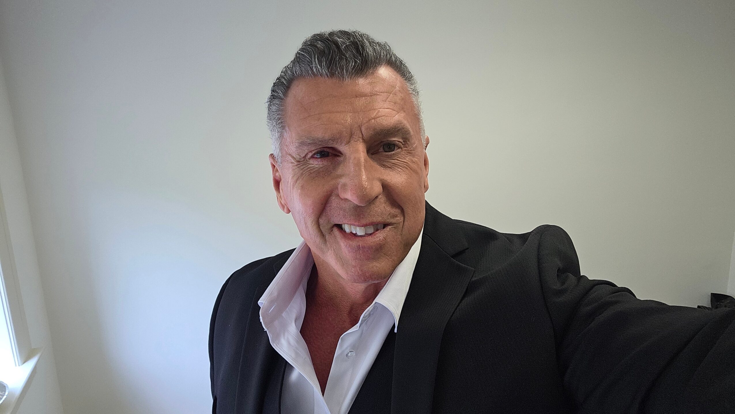 Man in a suit taking a selfie indoors, smiling, with a plain wall background.