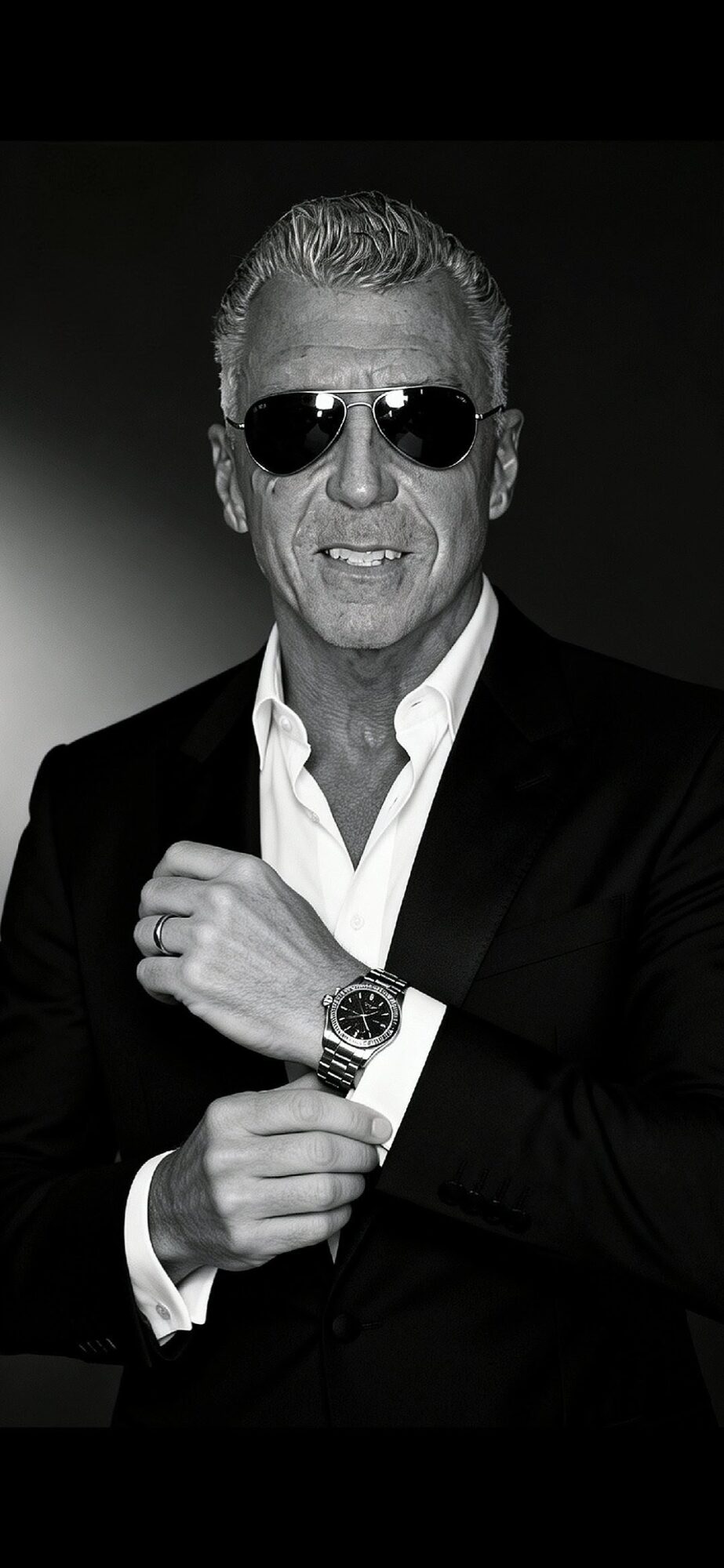 Man with sunglasses, wearing a suit, adjusting his watch, smiling, in black and white.