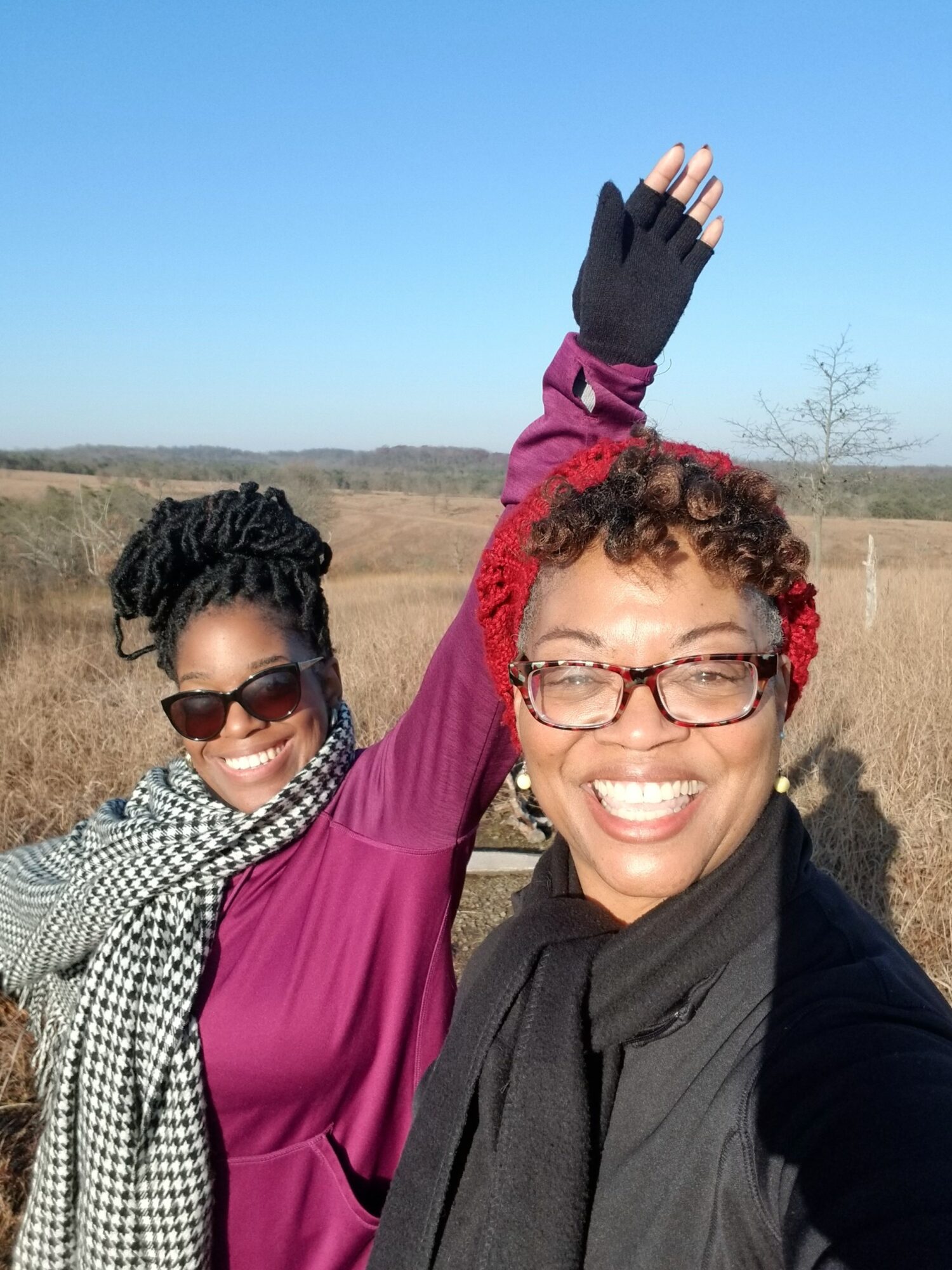 Two women outdoors, one with red hair and glasses, smiling and waving, the other with black hair and sunglasses, smiling.