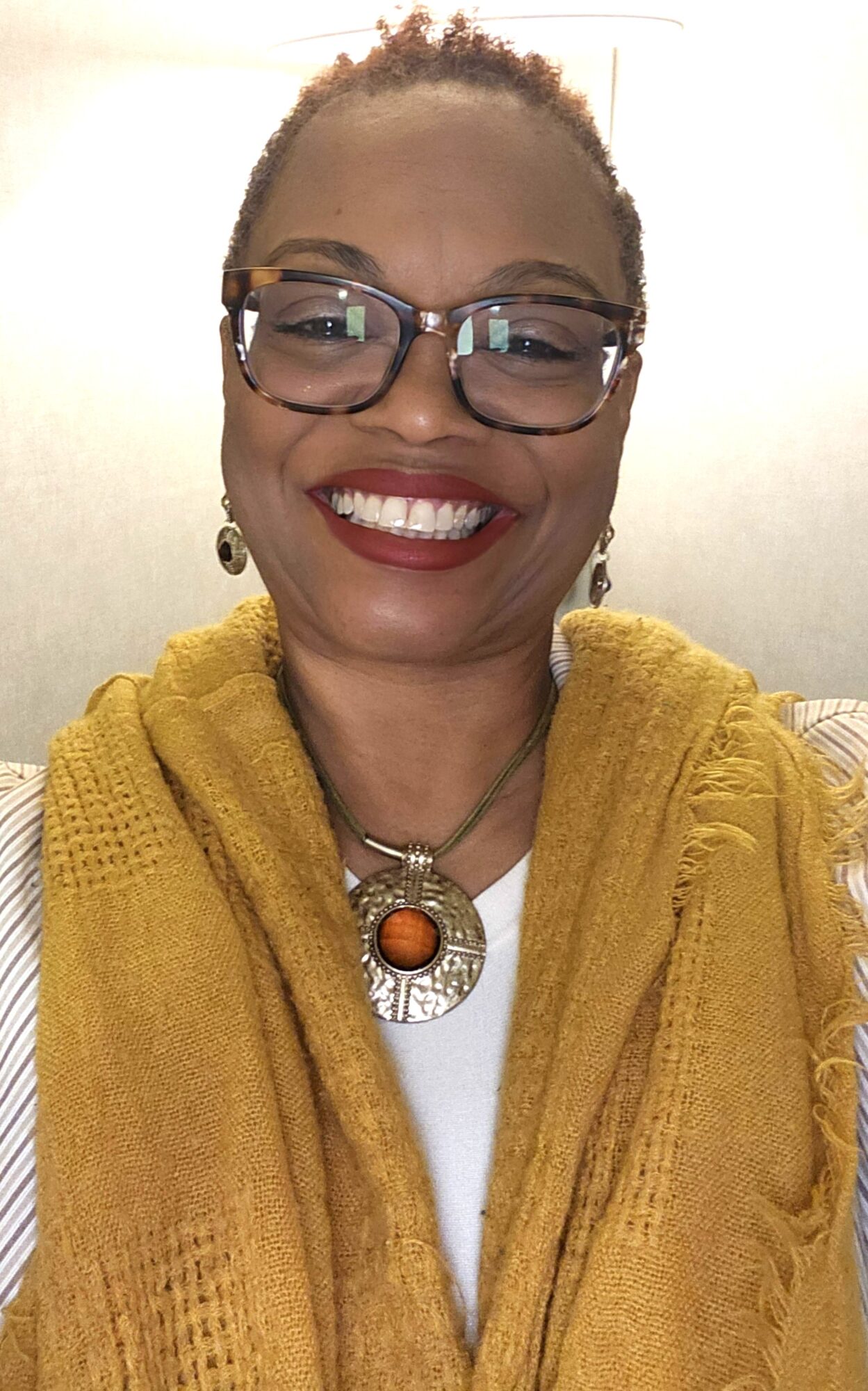 Smiling woman with glasses, earrings, and a large necklace, wearing a yellow scarf or shawl, against a plain background.