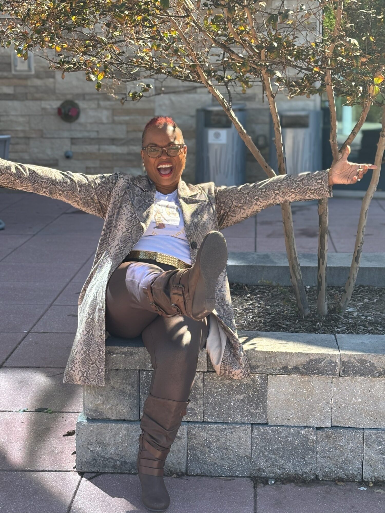 Person sitting on a concrete ledge with arms outstretched, smiling, outdoors with trees and trash bins behind.