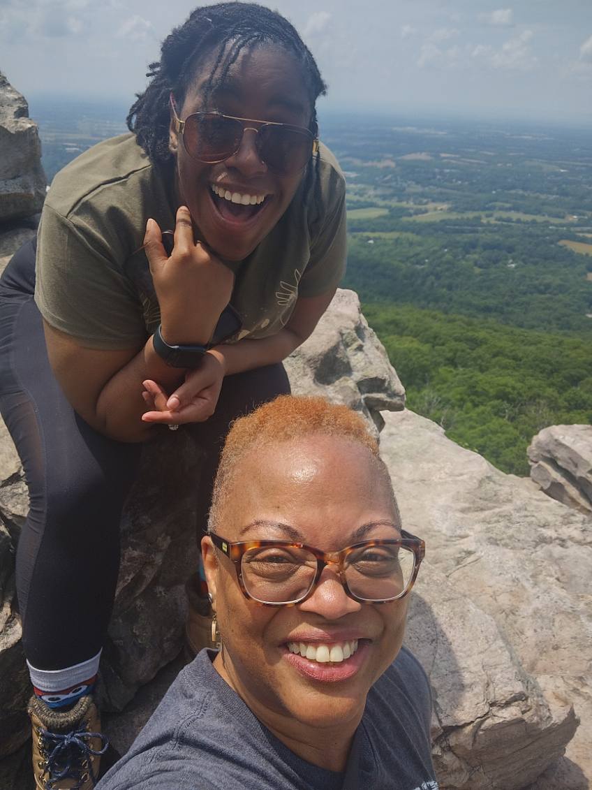 Two smiling women outdoors on a rocky hilltop with a landscape view in the background.
