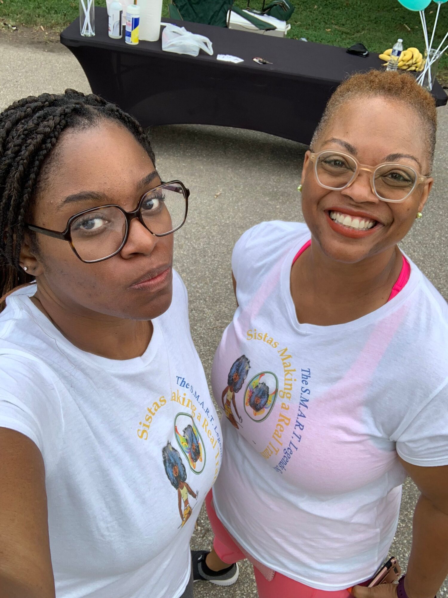 Two women wearing glasses and white T-shirts smiling outdoors, one with short hair and the other with braided hair.