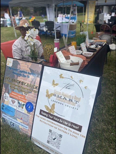 Outdoor market stall with a person behind a table displaying jewelry and promotional signs, under a blue canopy.
