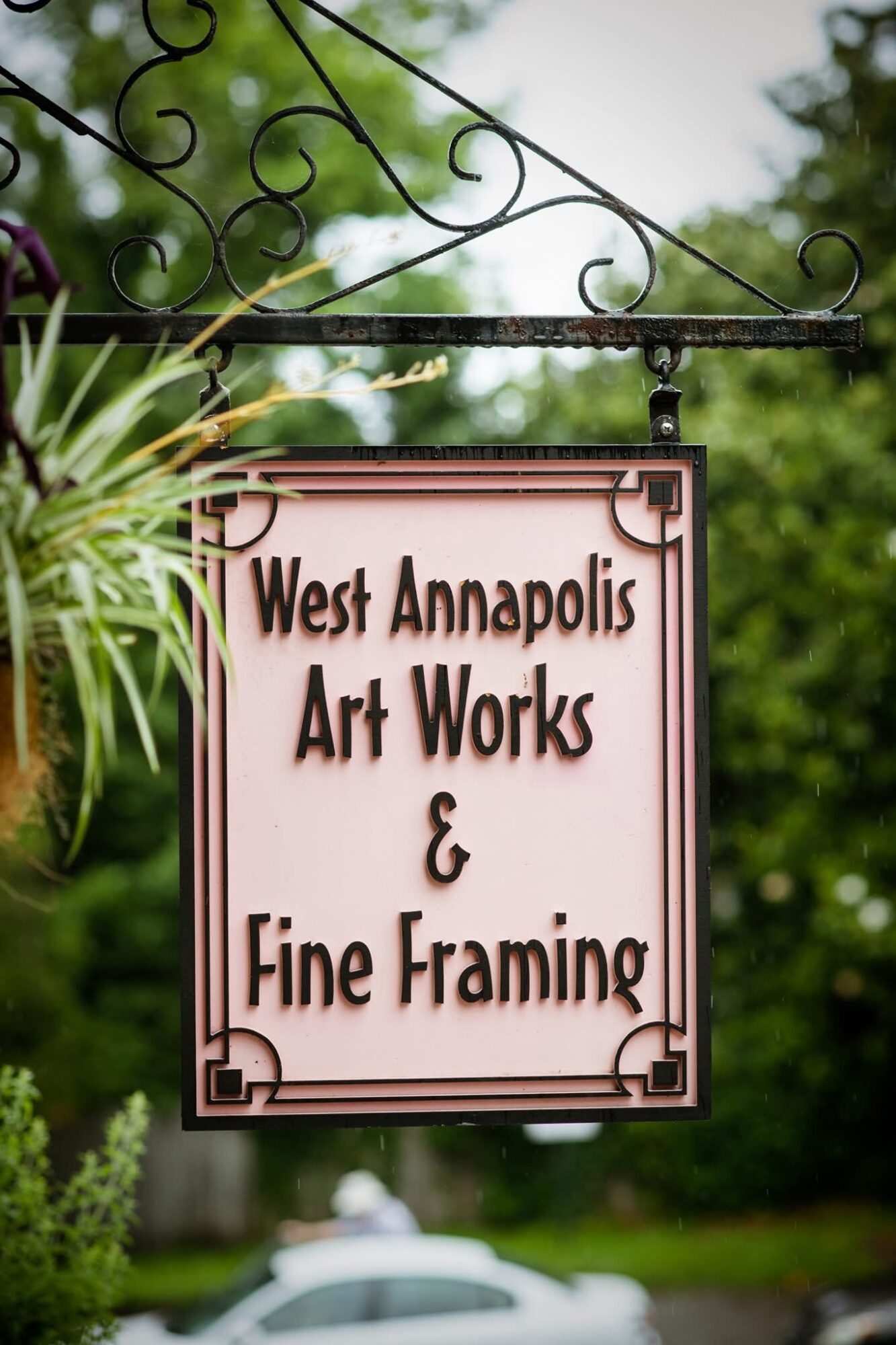 Sign for West Annapolis Art Works and Fine Framing hanging outdoors with greenery in background.