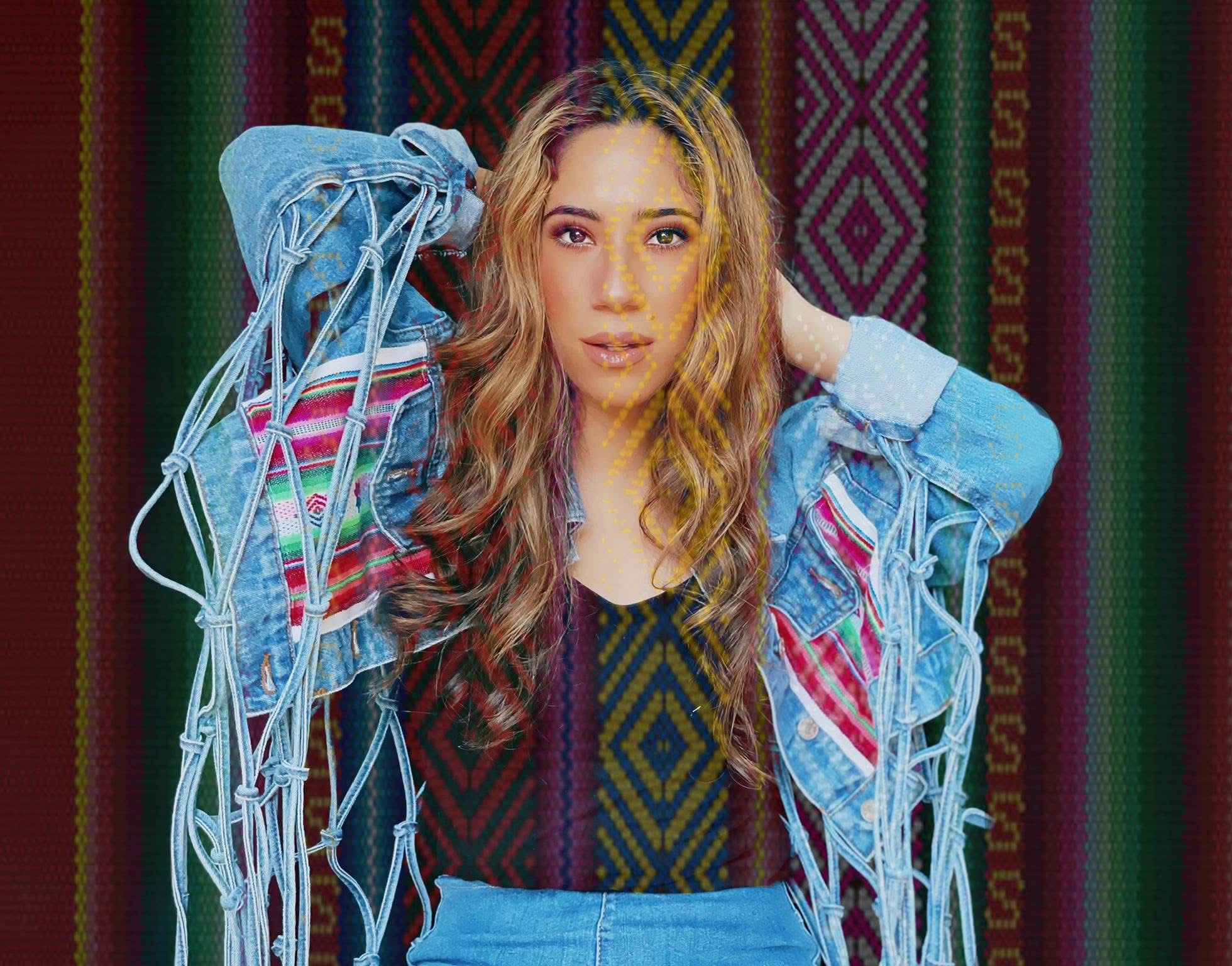 Woman with long wavy hair wearing a denim jacket over a patterned top, standing in front of colorful woven background.