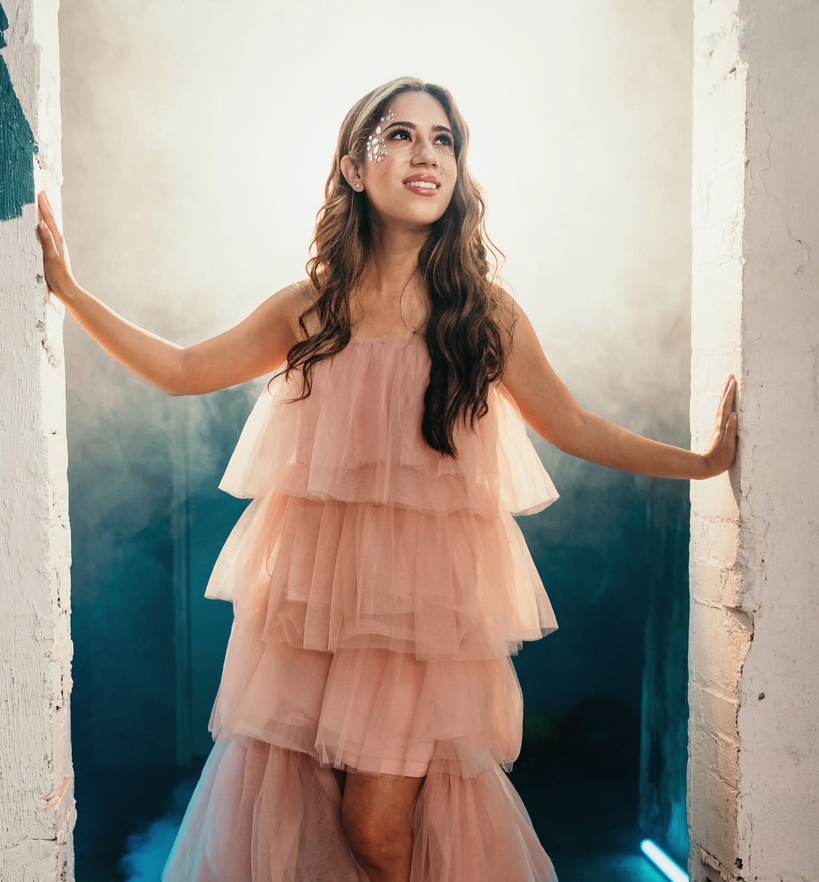 Young woman in a peach layered dress standing between two walls, smiling and looking upward.