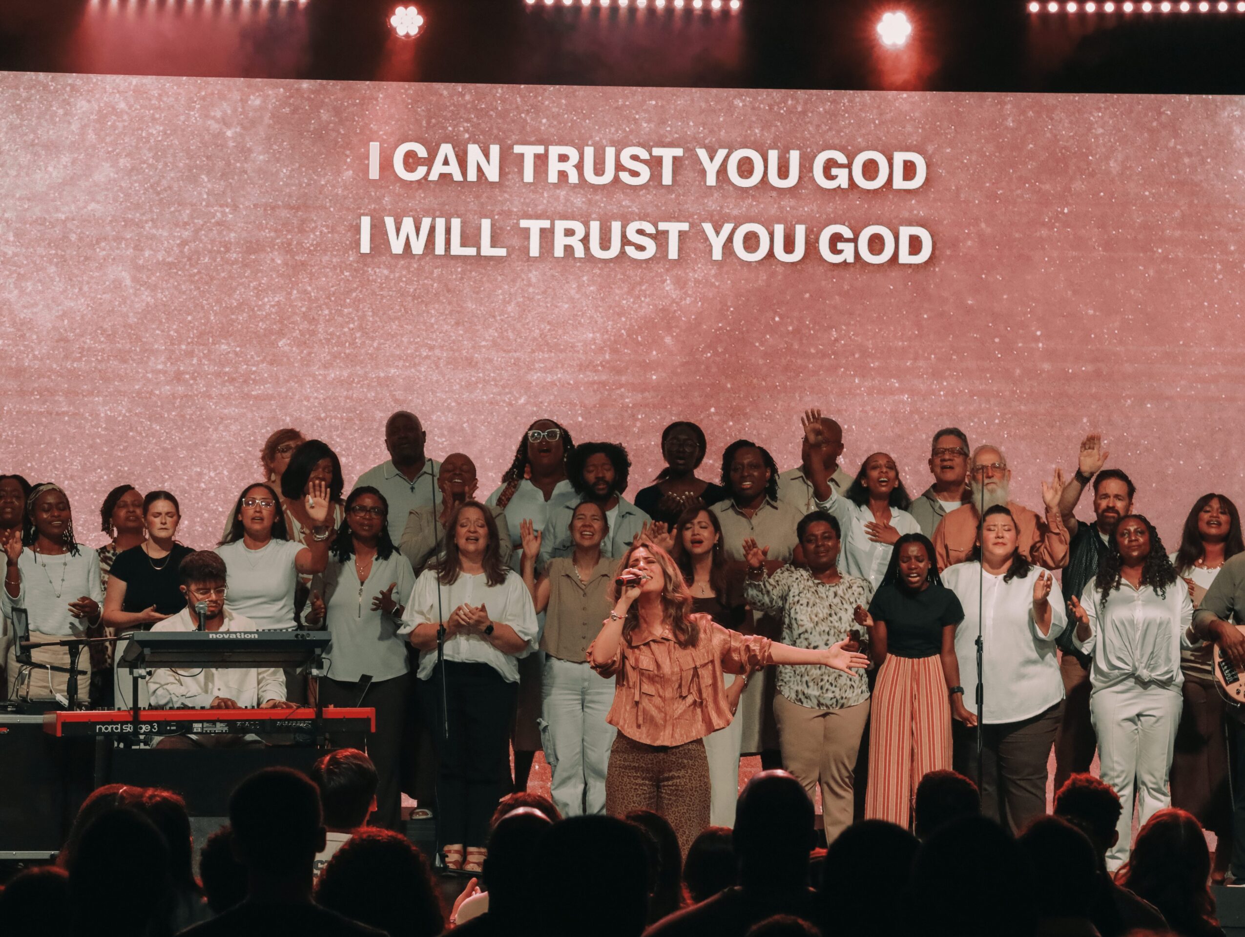 Group of people on stage singing with lyrics 'I CAN TRUST YOU GOD, I WILL TRUST YOU GOD' displayed behind them.