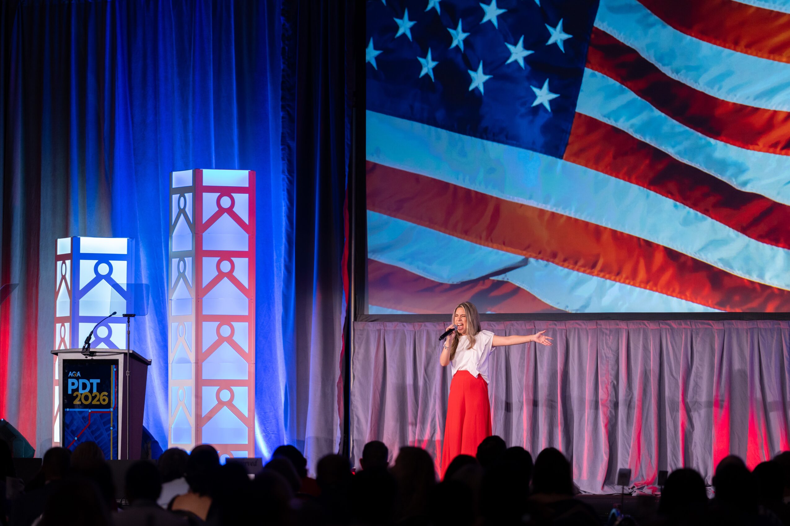 A woman on stage with a microphone, pointing, in front of a large American flag backdrop, with an audience watching.