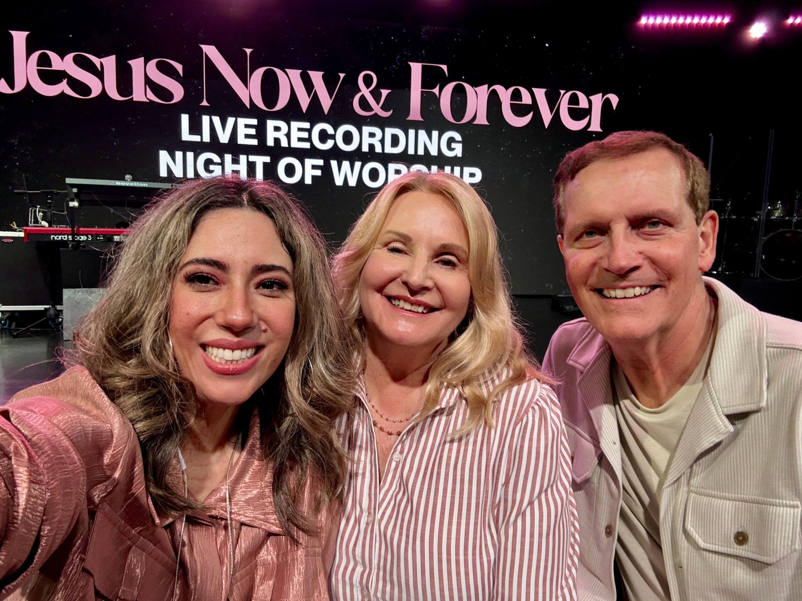 Three smiling people taking a selfie in front of a sign that reads 'Jesus Now & Forever' and 'Night of Worship'.