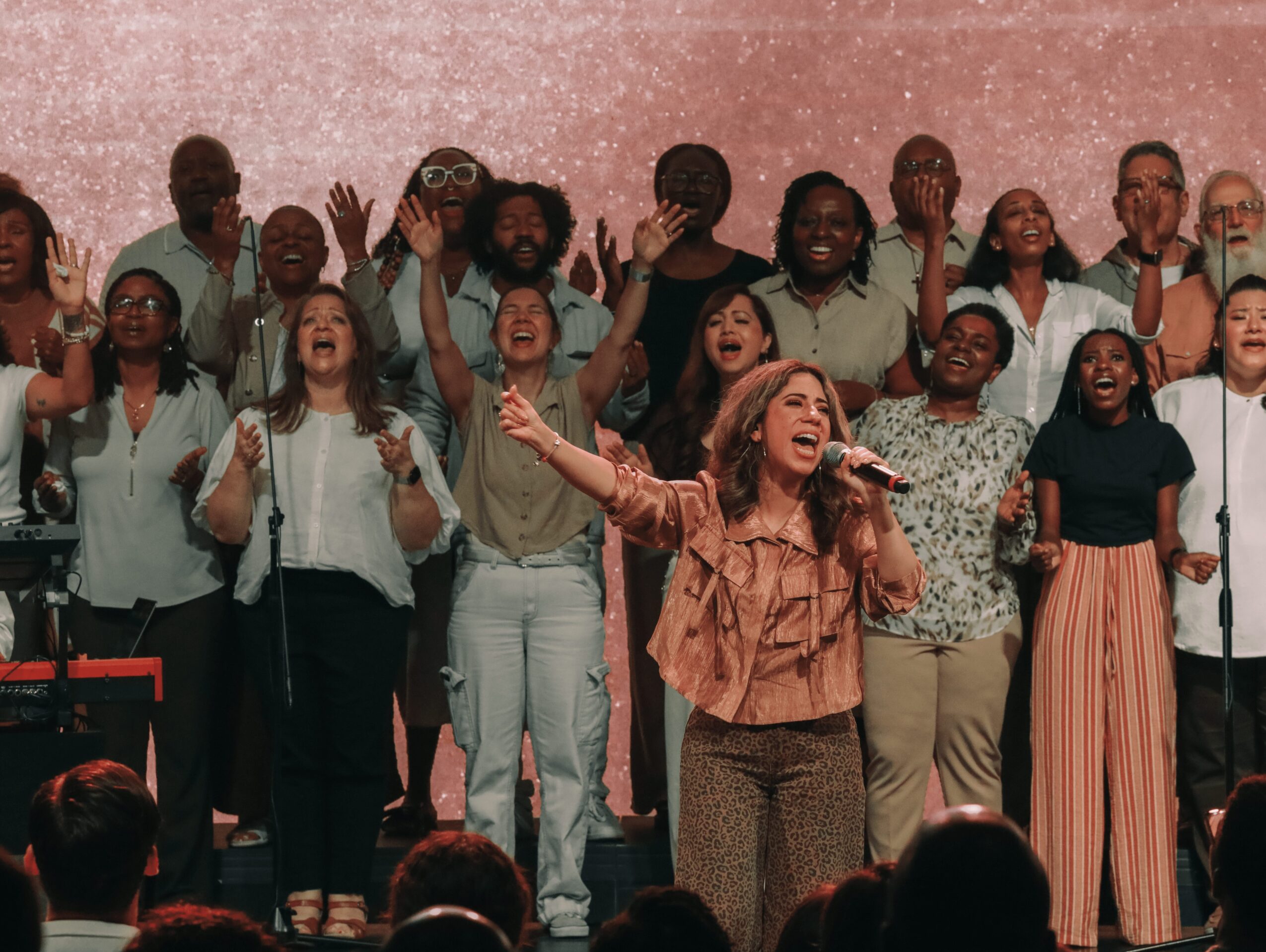 A woman sings into a microphone on stage with a diverse group of people behind her, raising their hands.
