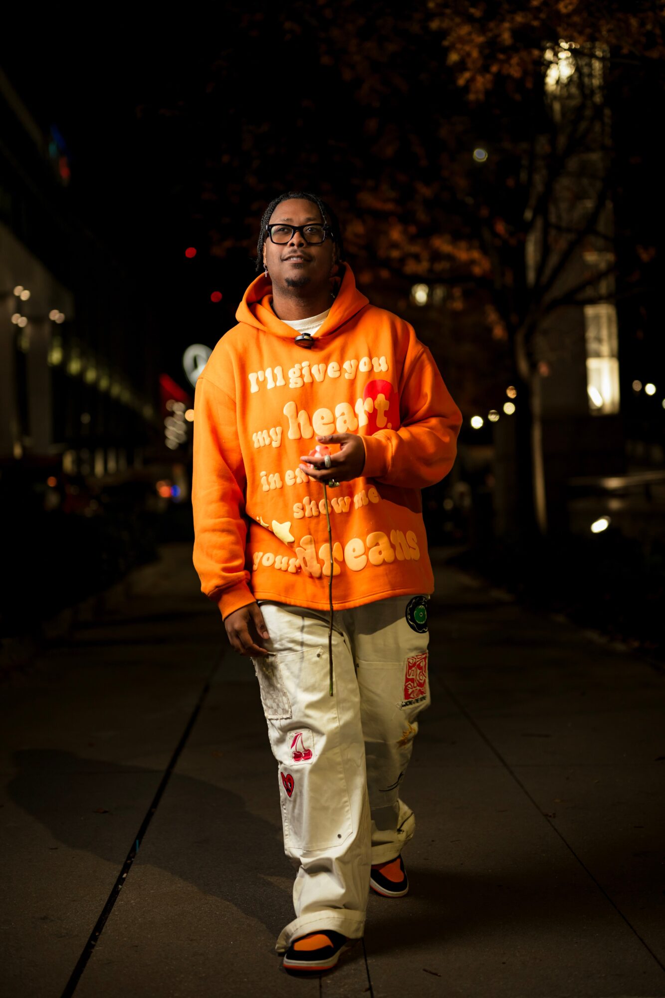 Person walking on a city street at night, wearing an orange hoodie and light-colored pants, holding a phone.