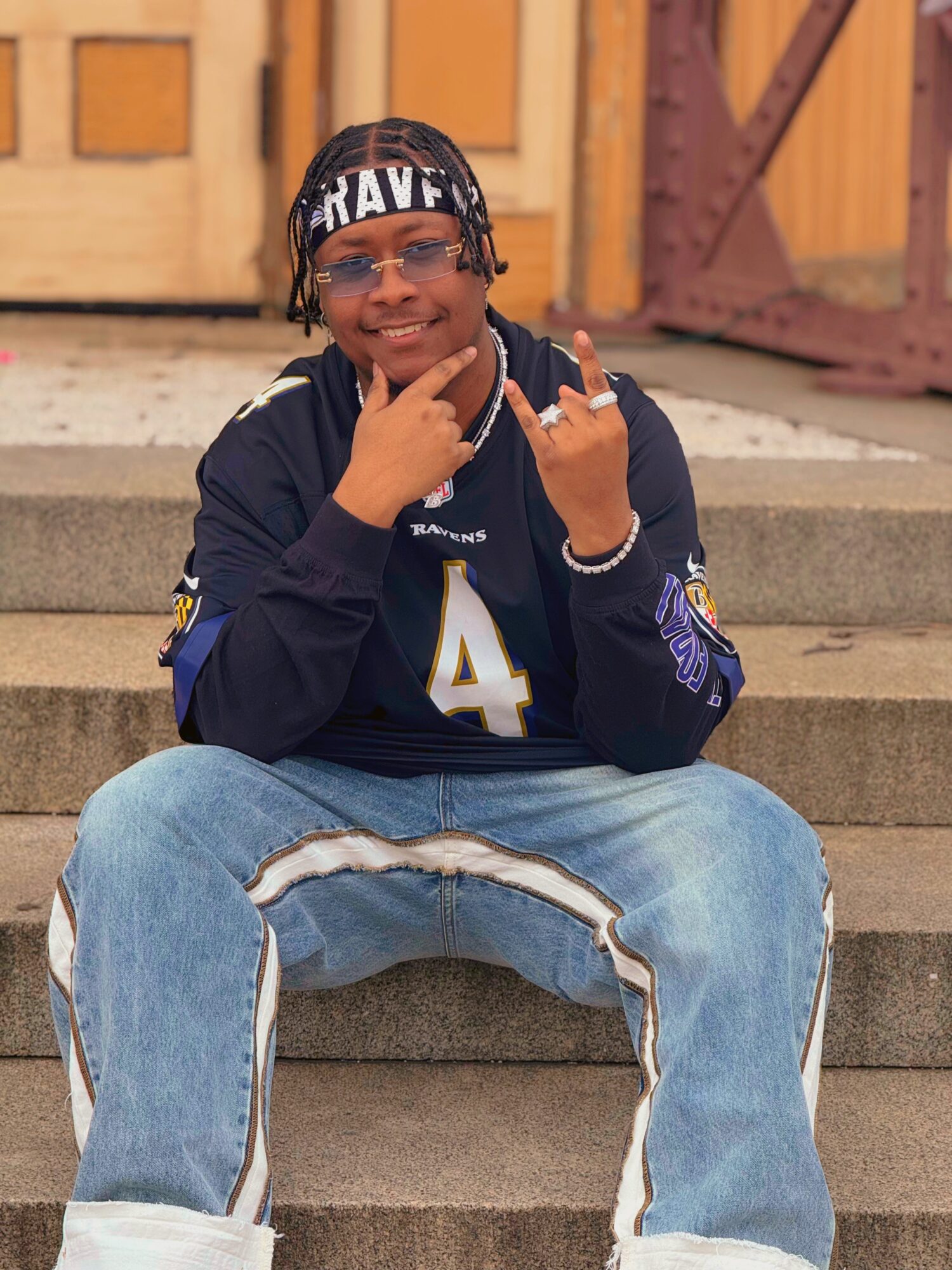 Young person sitting on steps, wearing glasses, a dark jacket, and jeans, smiling and making hand gestures.