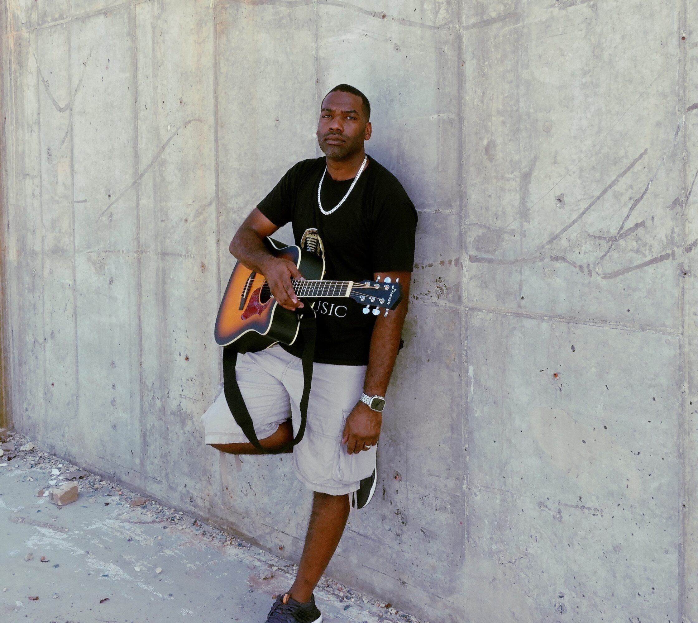 Young man with a guitar leaning against a concrete wall, wearing a black t-shirt, shorts, and a chain necklace.