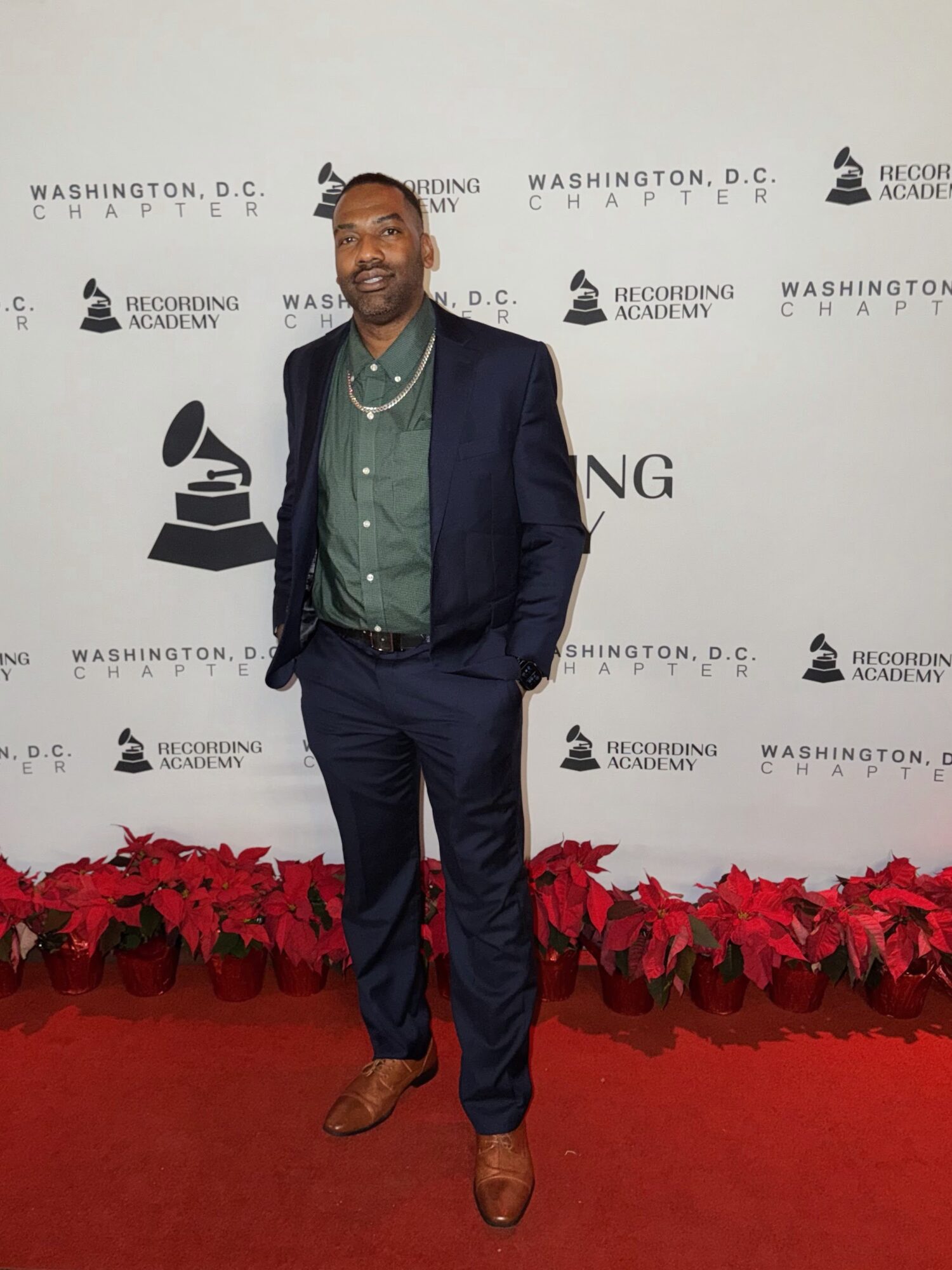 Man in a dark suit and green shirt standing on red carpet with poinsettia plants, smiling, backdrop with logos and text.