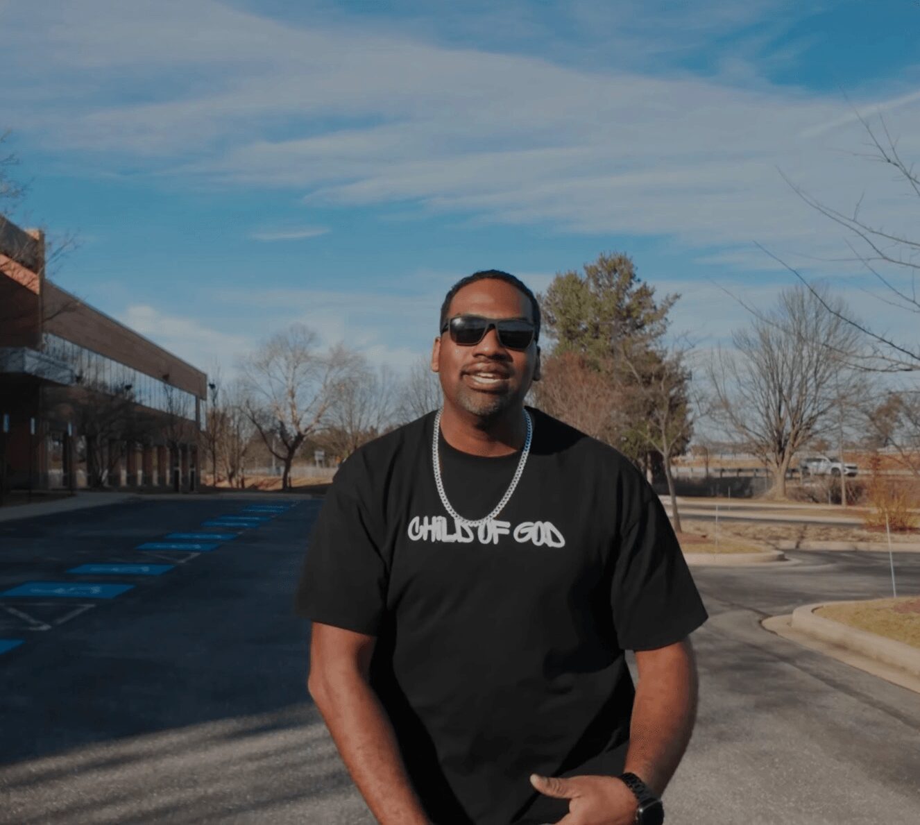 Man wearing sunglasses and a black T-shirt with text, standing outdoors on a sunny day.