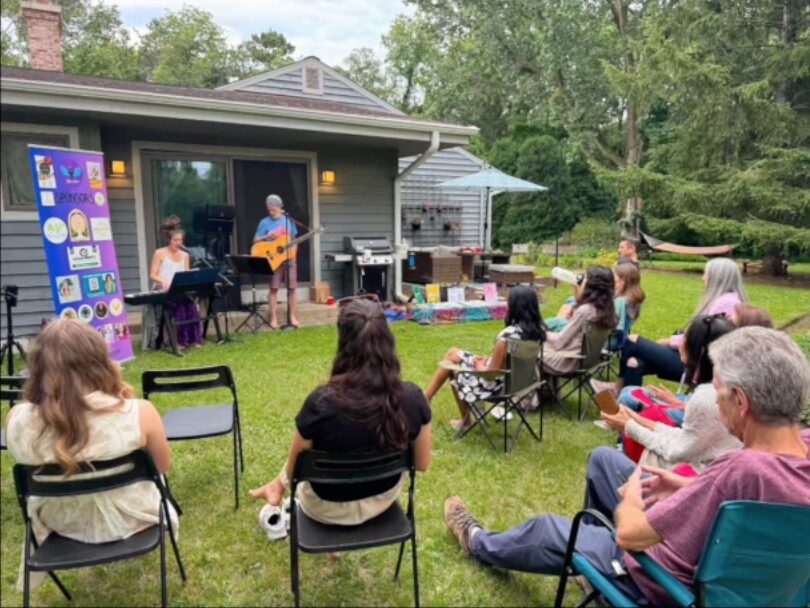 People sitting outdoors watching two musicians perform on a small stage, with trees and a house in the background.