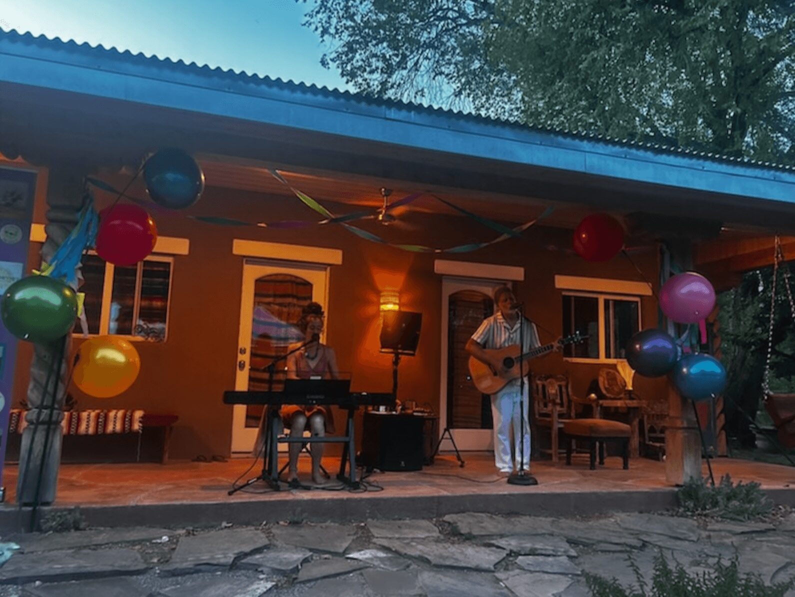 People performing music on a porch decorated with colorful balloons and streamers, with a house in the background.