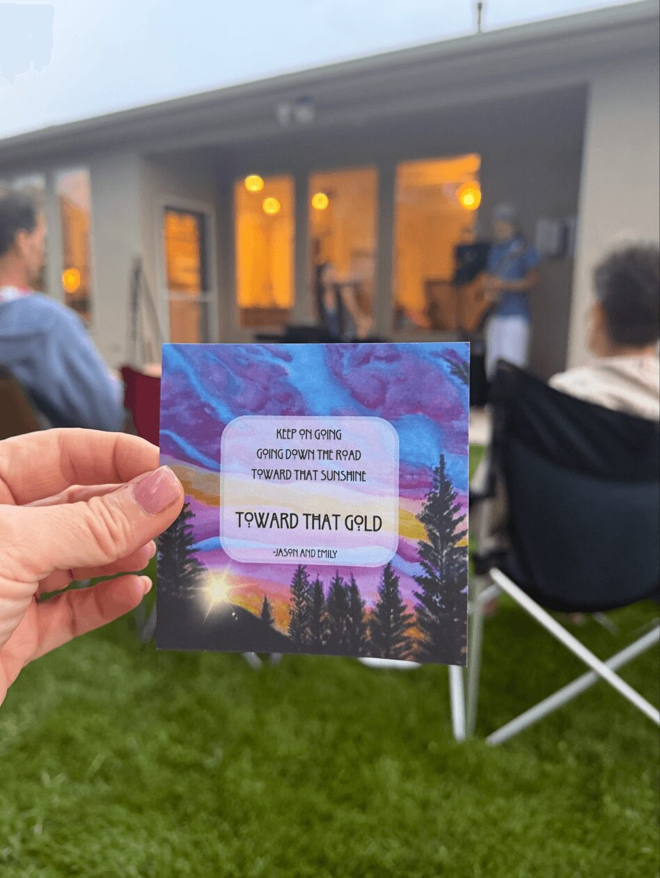 Person holding a colorful card with text, outdoors with people sitting on chairs and a building in the background.