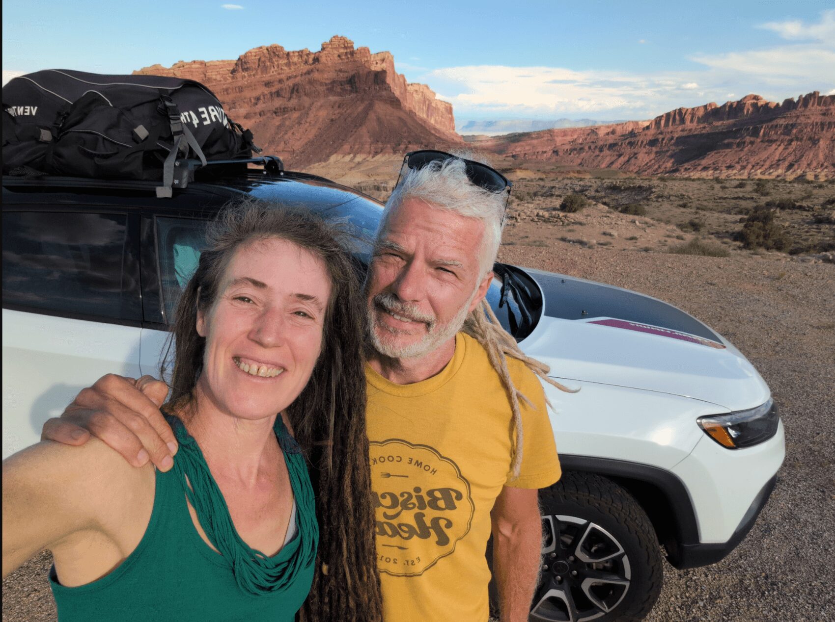 Two people smiling in front of a white vehicle with a desert landscape and red rock formations in the background.