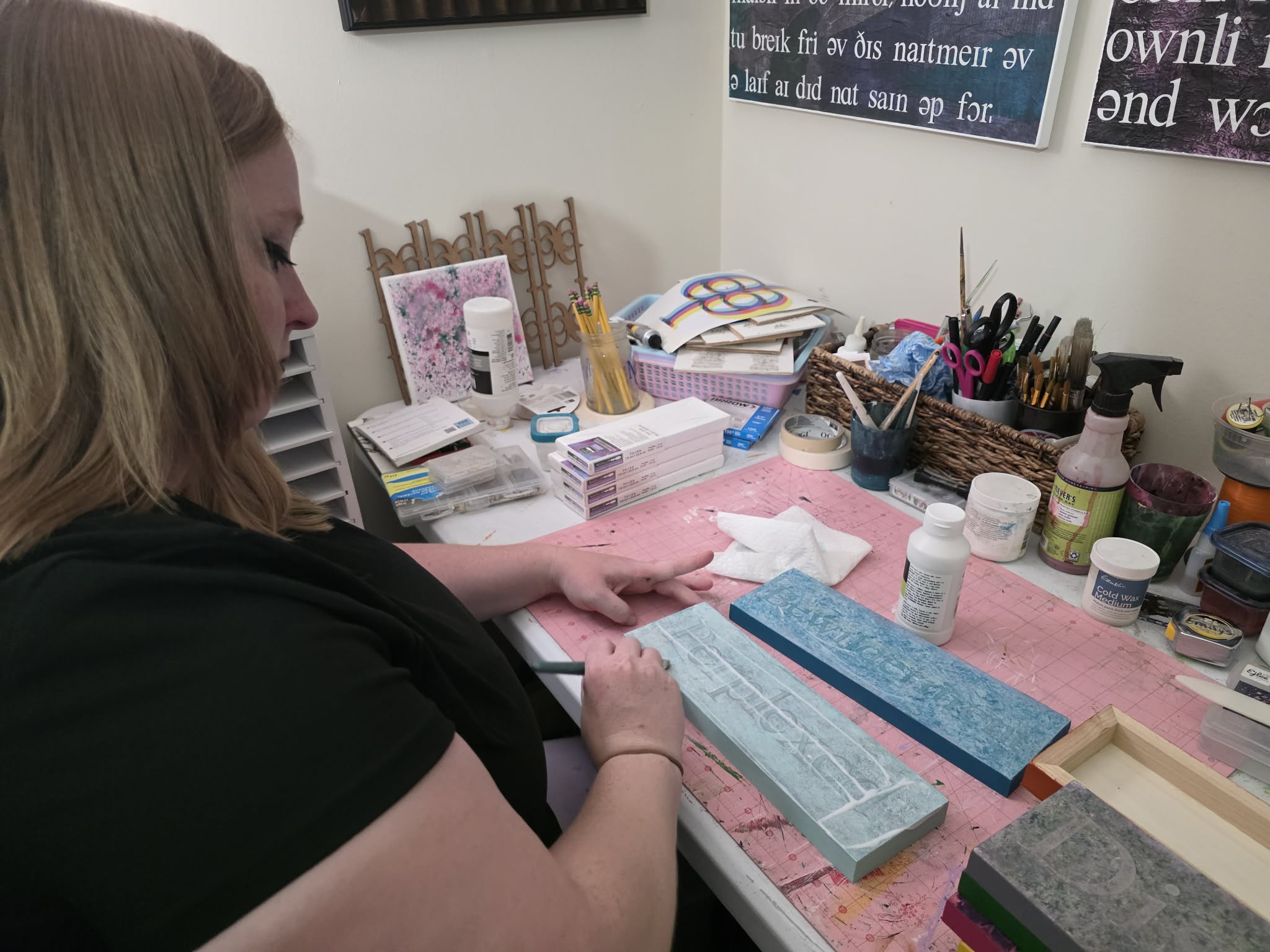 Woman sitting at a cluttered desk with various art supplies and papers, focusing on her work.