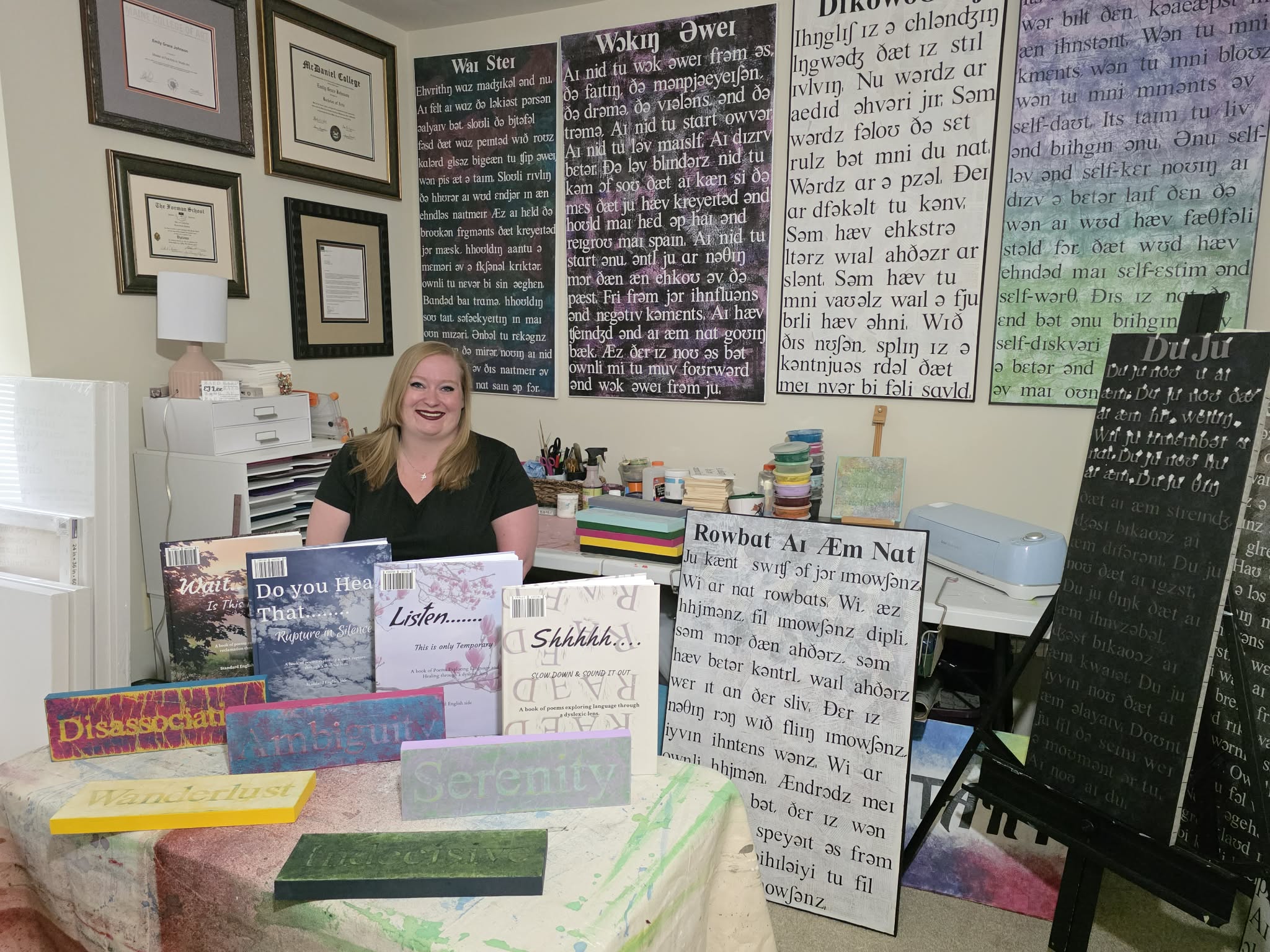Woman sitting at a desk surrounded by books and posters, smiling in a room with framed certificates on the wall.