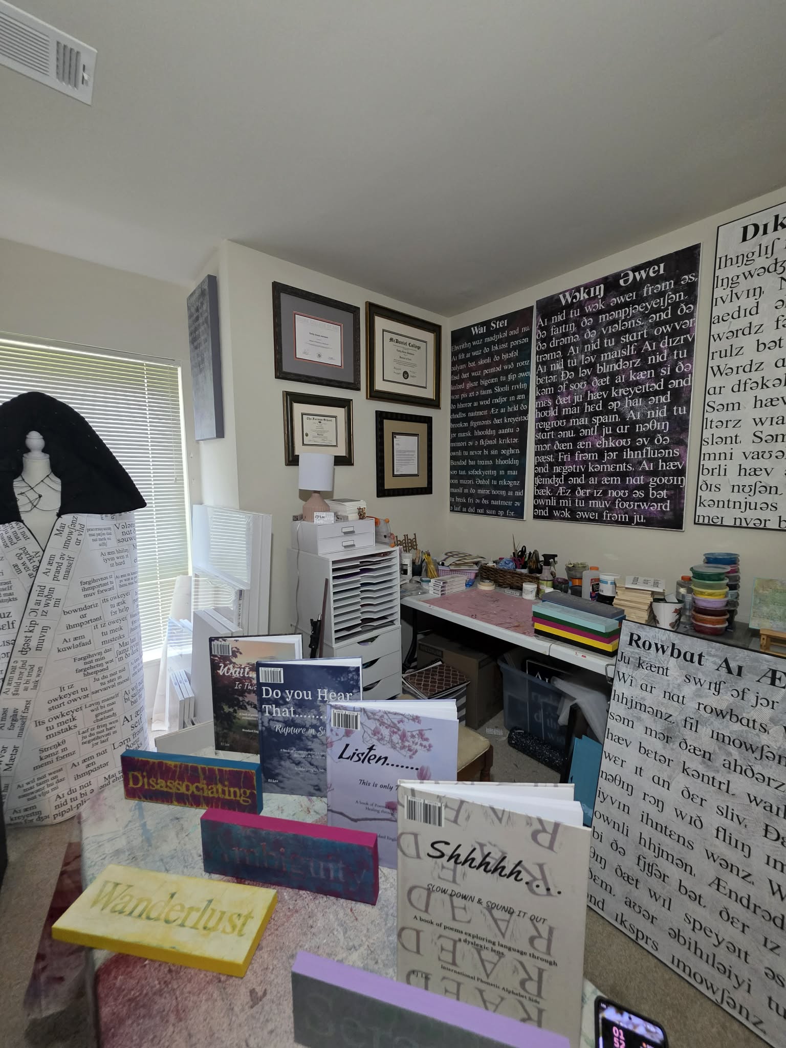 Room with framed certificates, chalkboard, and desk with books and papers, cluttered workspace, and a window with blinds.