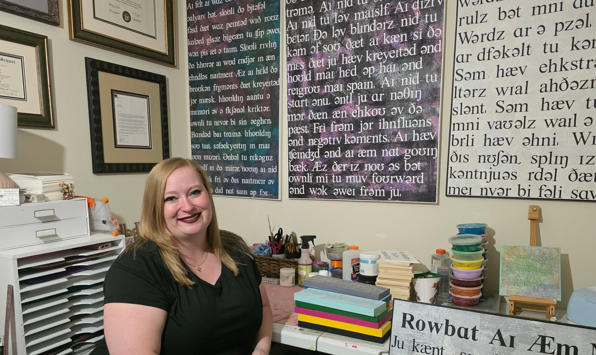 Smiling woman sitting at a desk with books, papers, and colorful containers in a classroom or office setting.